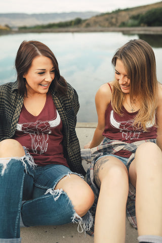 Two women sitting in front of small lake/pond wearing graphic tank tops with a steer skull design on the front. Also wearing flannel shirts, jeans, and converse and birkenstock sandals. PNW Style. Pacific Northwest. Wenatchee, Washington. Mountain Style. Golden Hour.