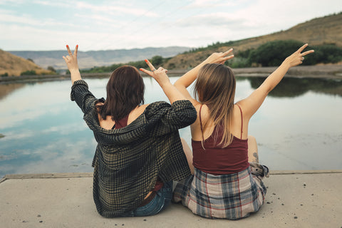 Two women sitting in front of small lake/pond wearing graphic tank tops with a steer skull design on the front. Also wearing flannel shirts, jeans, and converse and birkenstock sandals. PNW Style. Pacific Northwest. Wenatchee, Washington. Mountain Style. Golden Hour.