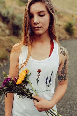 Woman standing in front of a dirt road wearing a white tank top featuring an illustration of Pacific Northwest Wildflowers. She is holding a bouquet of flowers. Jeans shorts. Summer time. Flowers. Floral. Wildflower. Wenatchee, Washington. PNW. Pacific Northwest Style.