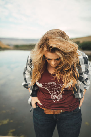 Woman standing in front of small lake/pond in Wenatchee, Washington. She is wearing a maroon colored tank top with a steer skull design on the front and jeans with a brown belt, and a flannel shirt. PNW. Pacific Northwest Style.