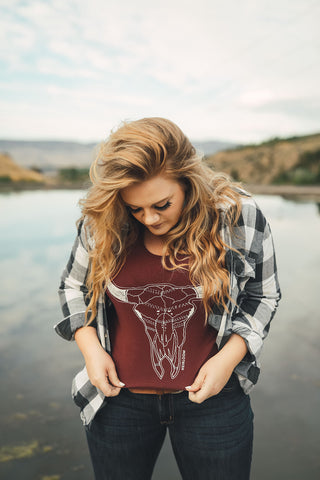 Woman standing in front of small lake/pond in Wenatchee, Washington. She is wearing a maroon colored tank top with a steer skull design on the front and jeans with a brown belt, and a flannel shirt. PNW. Pacific Northwest Style.