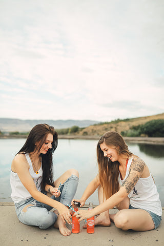 Two women sitting in front of small lake/pond opening up glass soda bottles. Wearing white tank tops with jeans and jeans shorts. Summer time. Wenatchee, Washington. PNW Style. Pacific Northwest. Mountains. Mountain Style.