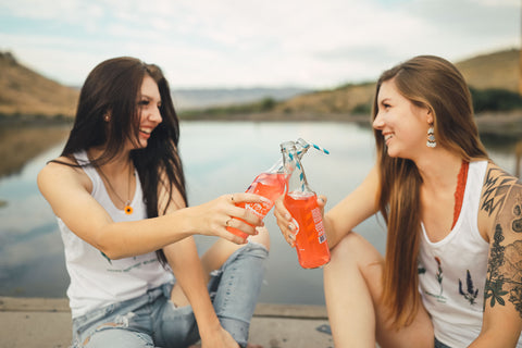 Two women sitting in front of small lake/pond opening up glass soda bottles. Wearing white tank tops with jeans and jeans shorts. Summer time. Wenatchee, Washington. PNW Style. Pacific Northwest. Mountains. Mountain Style.