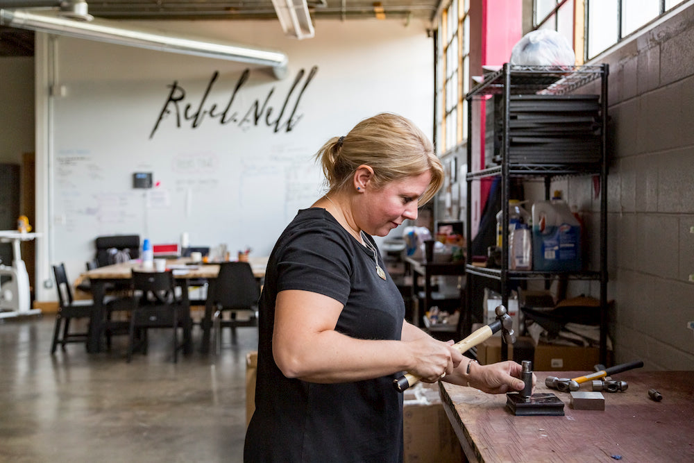 Rebel Nell co-founder Amy Peterson works with a hammer and jewelry-making tools at a worktable in a light-filled studio. 
