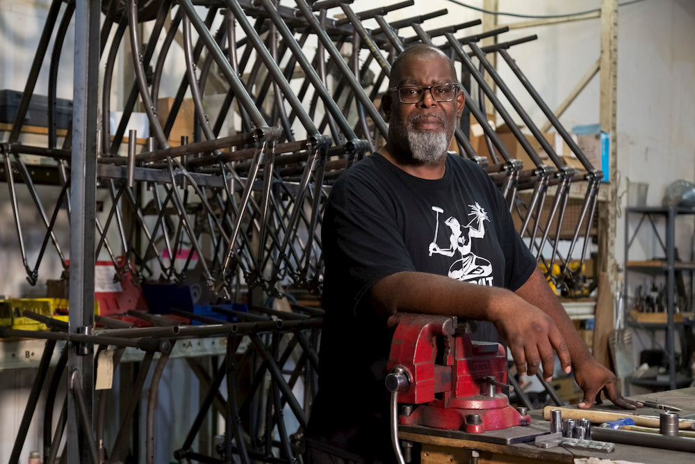 Detroit Bikes employee and bike mechanic Henry Ford II leans on a work table in a factory with bike frames lining the wall behind him.