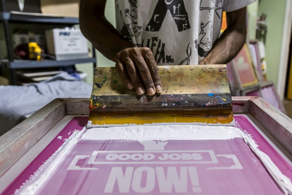Detail of a screen printer’s hands applying ink to a screen that says “GOOD JOBS NOW!”