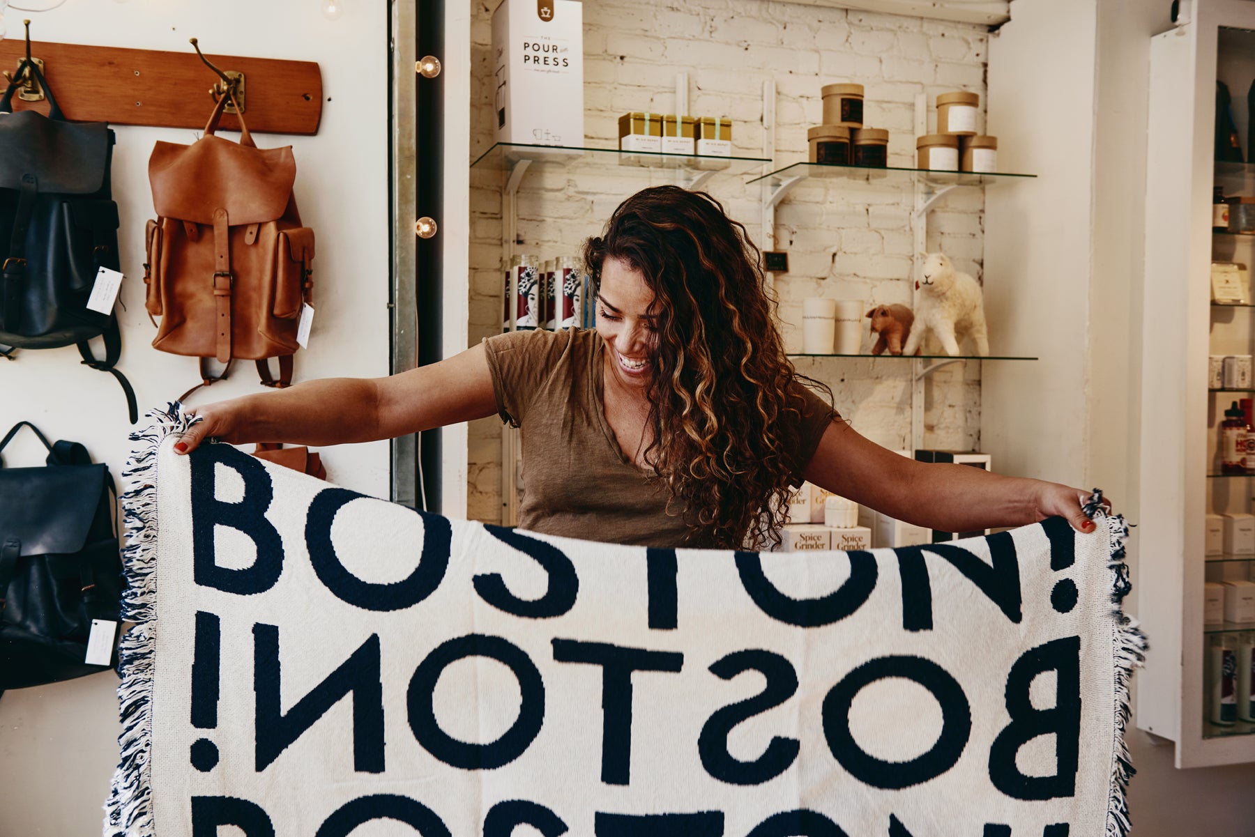 Sofi Madison stands in her Olives & Grace boutique holding up a blanket printed with the word “BOSTON!” in bold black type.
