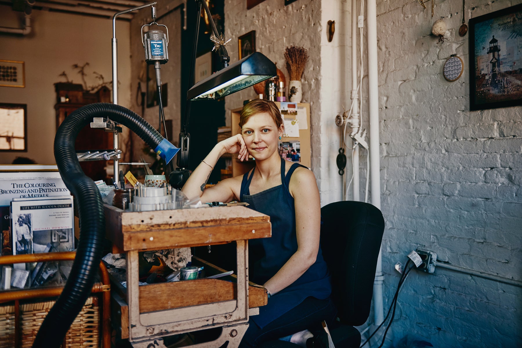 A portrait of Hannah Blount wearing an apron and sitting at a workbench in her jewelry studio against a white brick wall. 