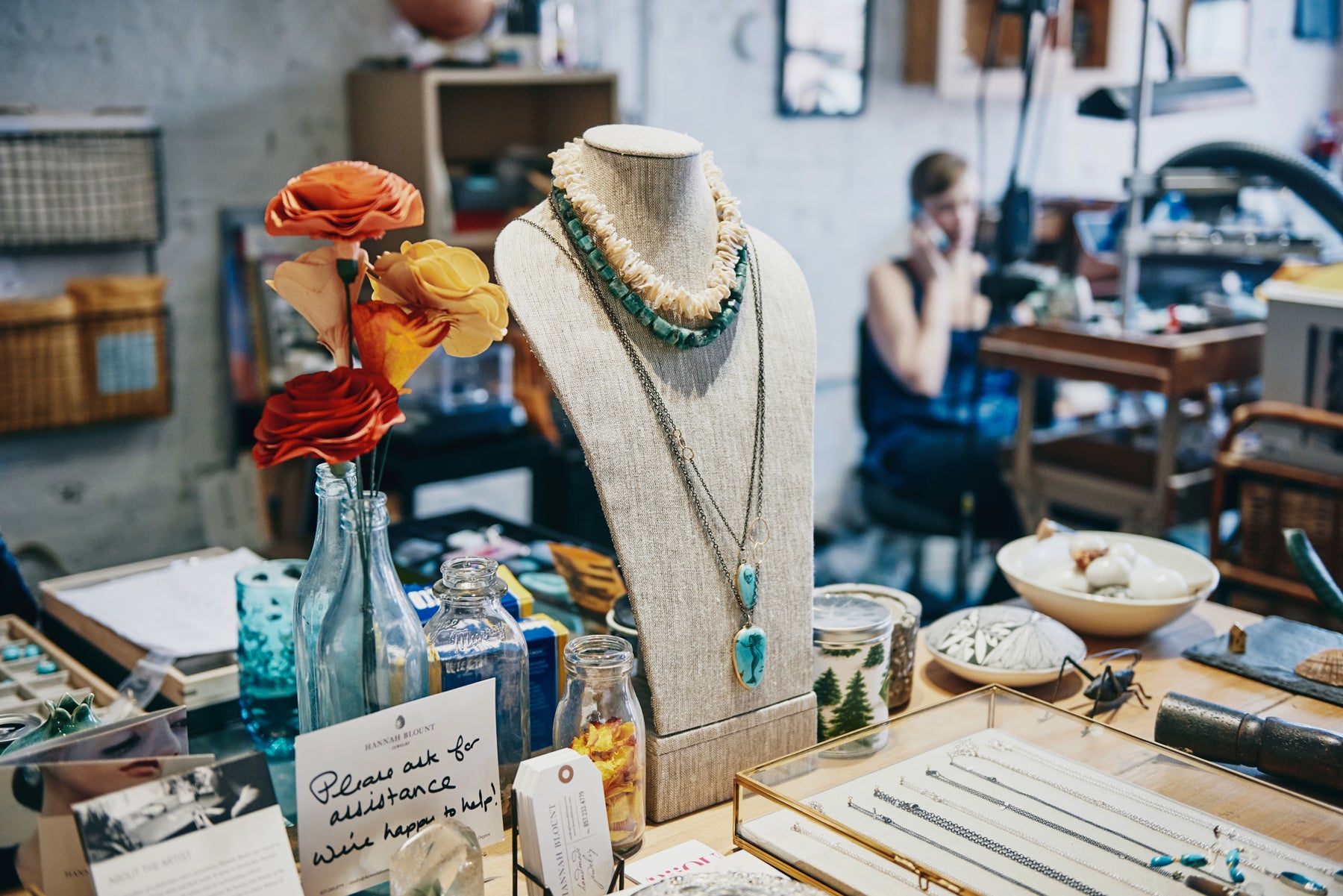 Image of a jewelry display featuring necklaces, a vase of flowers, and a handwritten note that says, “Please ask for assistance. We’re happy to help!” Hannah Blount talks on the phone in the background.