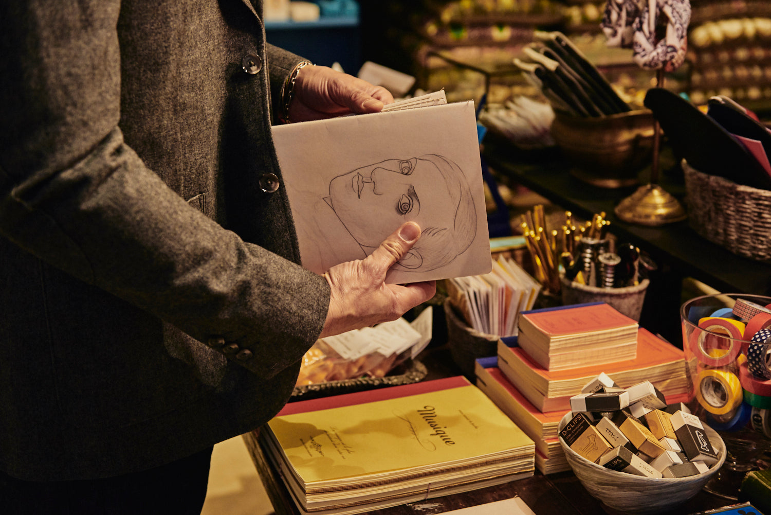 Detail of a man’s hands holding a pencil drawing of a face. A nearby table is merchandised with books, washi tape, paperclips, and décor items.