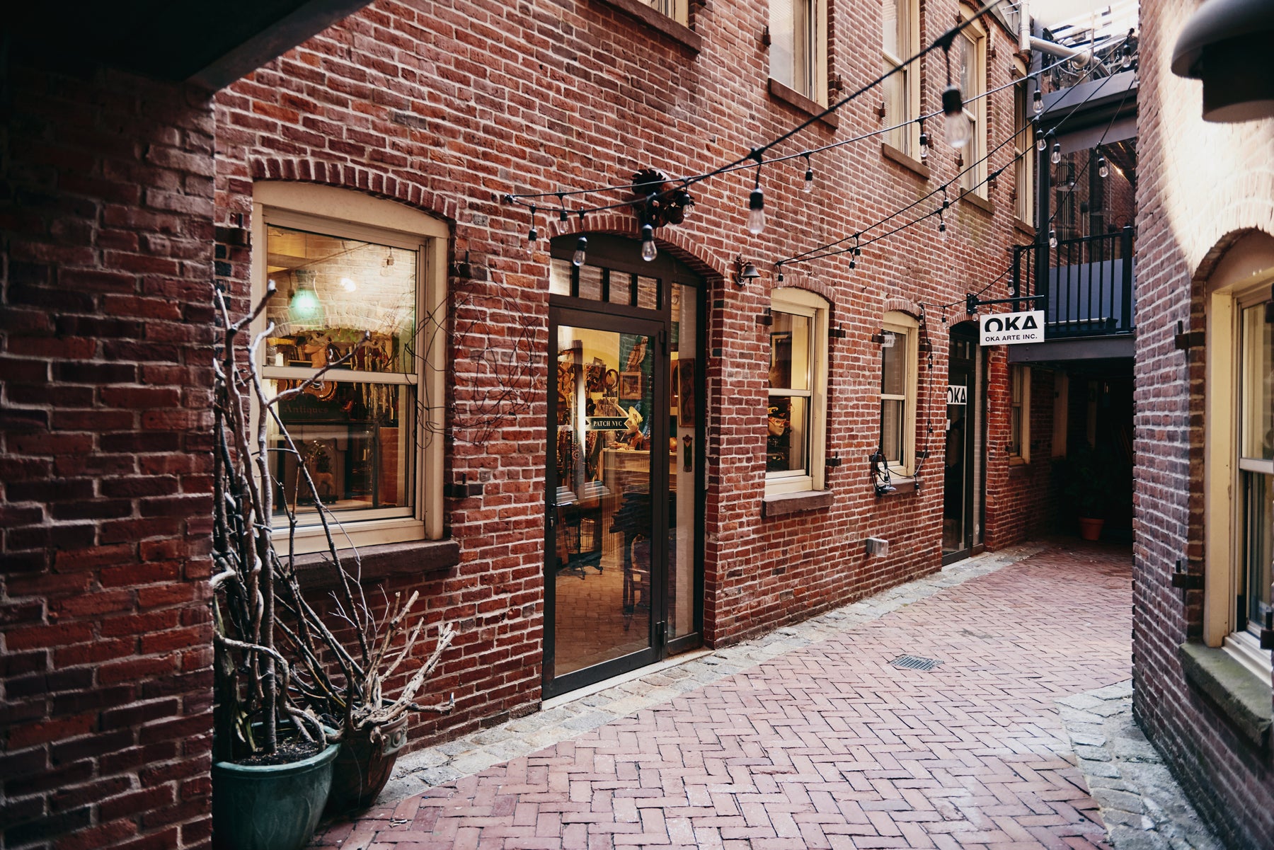 Image of the exterior of a brick retail building in an alley in Boston’s South End neighborhood. Two store signs say, “PATCH NYC” and “OKA ARTE INC.” String lights crisscross the narrow space between buildings.