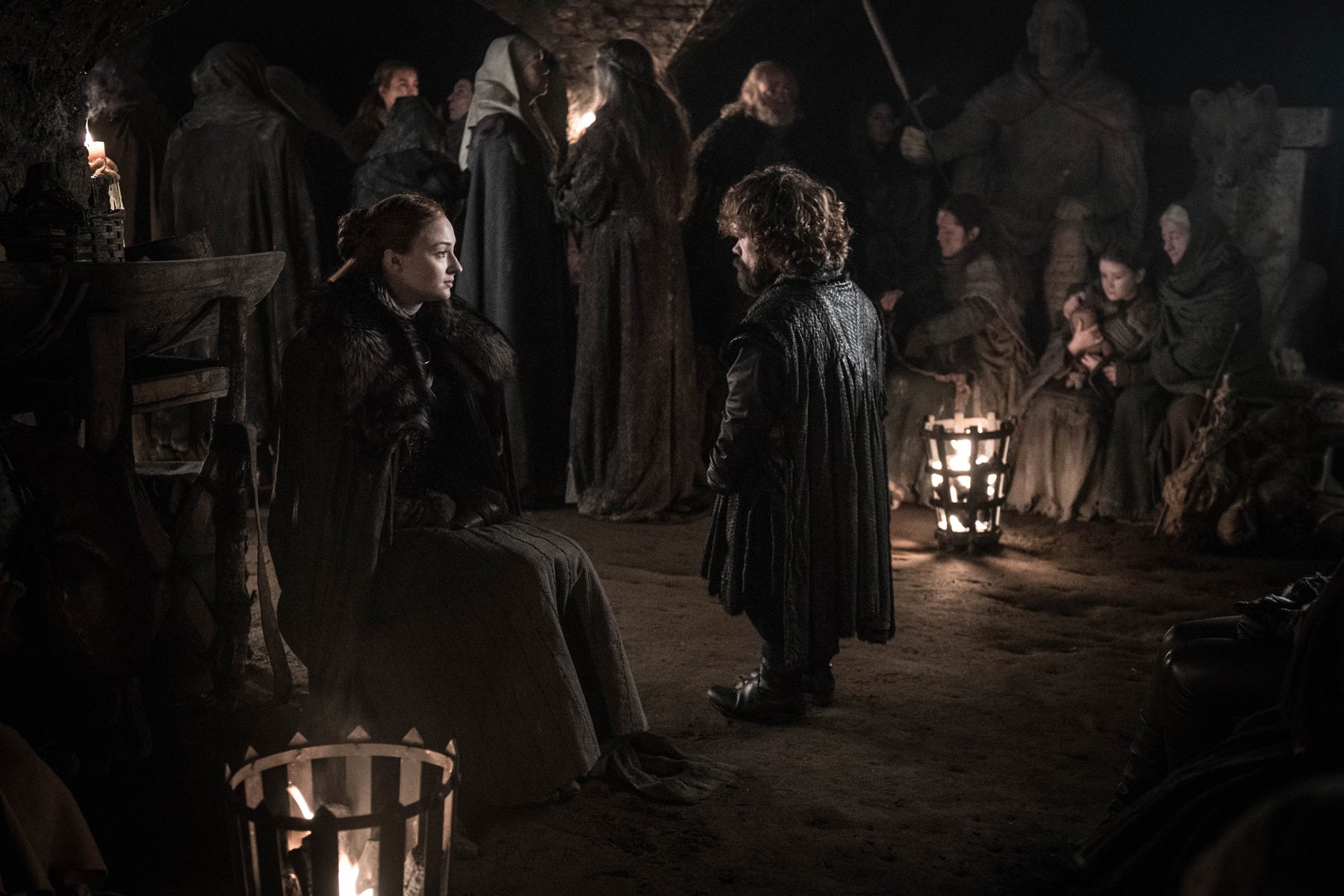 Sansa Stark is seated and facing Tyrion Lannister in the crypts of Winterfell, where women and children sit and stand in the background.
