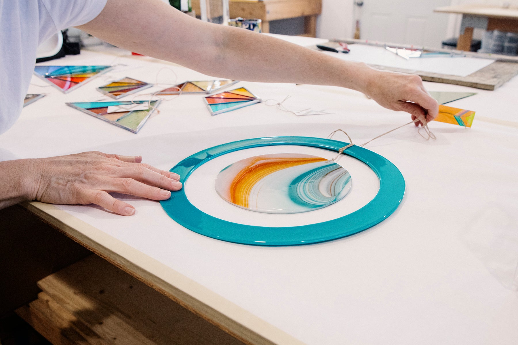 Detail photograph of Debbie Bean laying down a round, pale blue piece of stained glass with a string on top, in her studio.