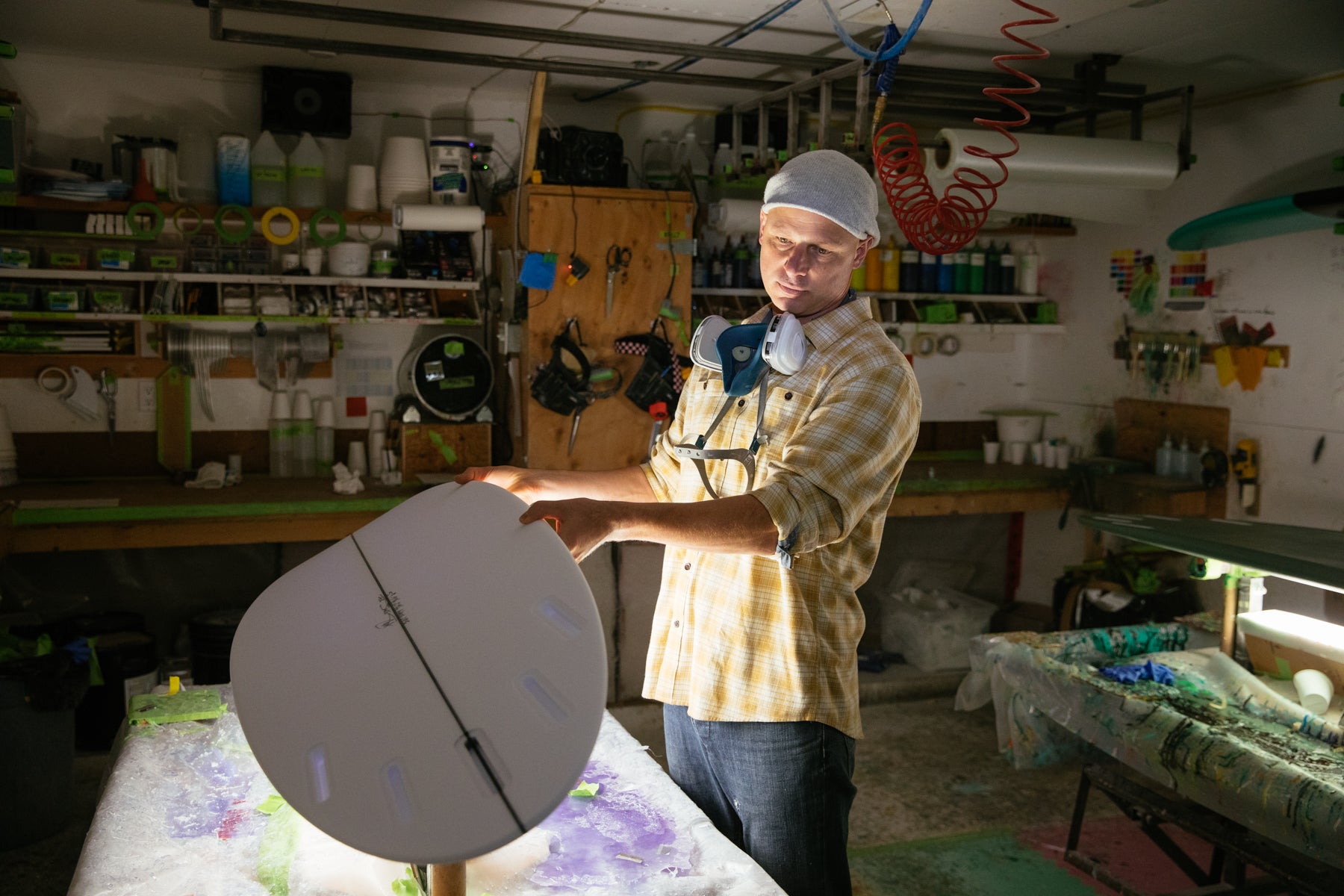 Stef Aftanas in yellow checkered shirt and jeans, inspects a surfboard in his workshop.