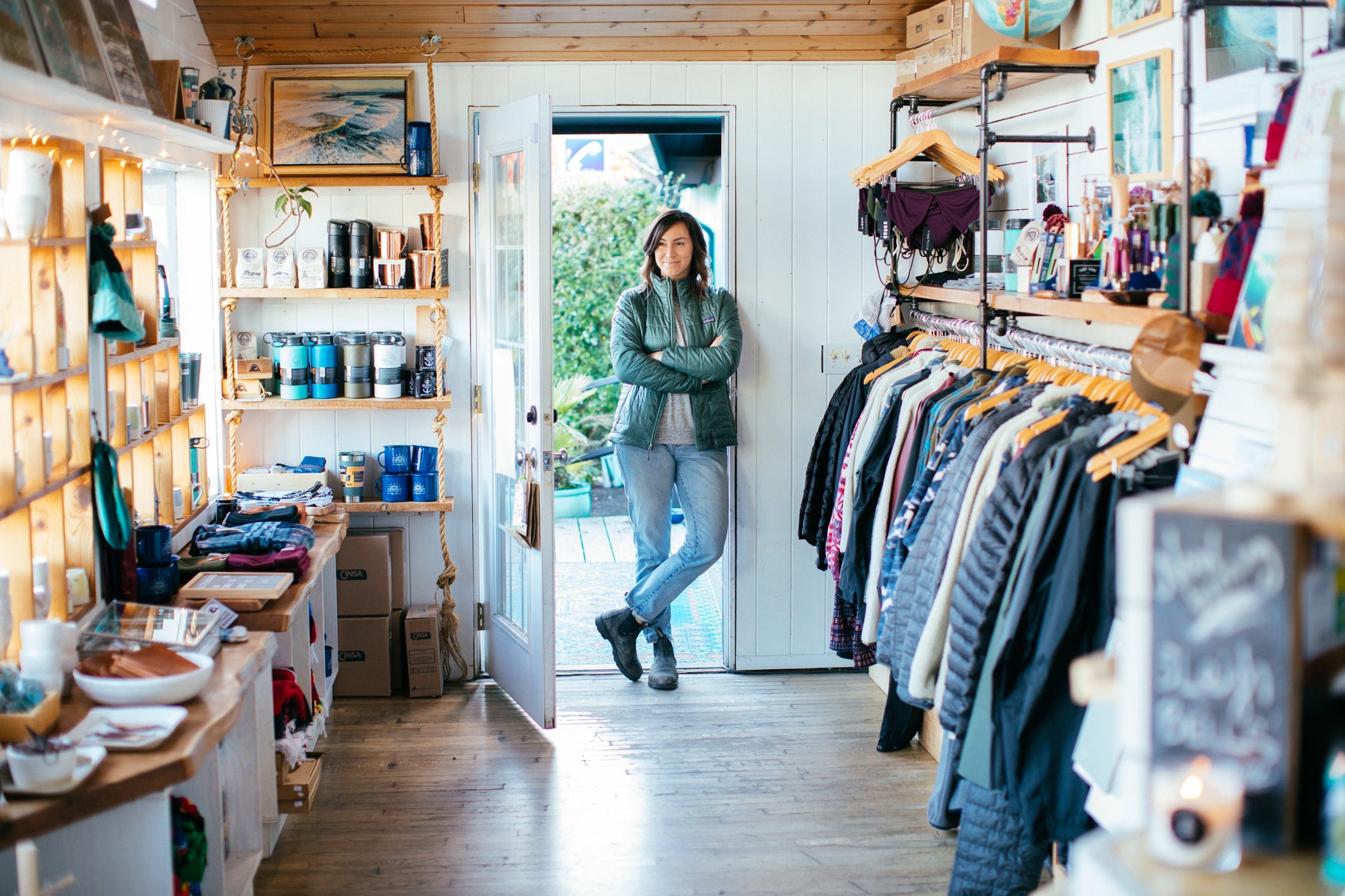 Jen Thorpe in green jacket and jeans, stands at the door of her store, Caravan Beach Shop. She overlooks the collection of homegoods on the left and clothing on the right. 