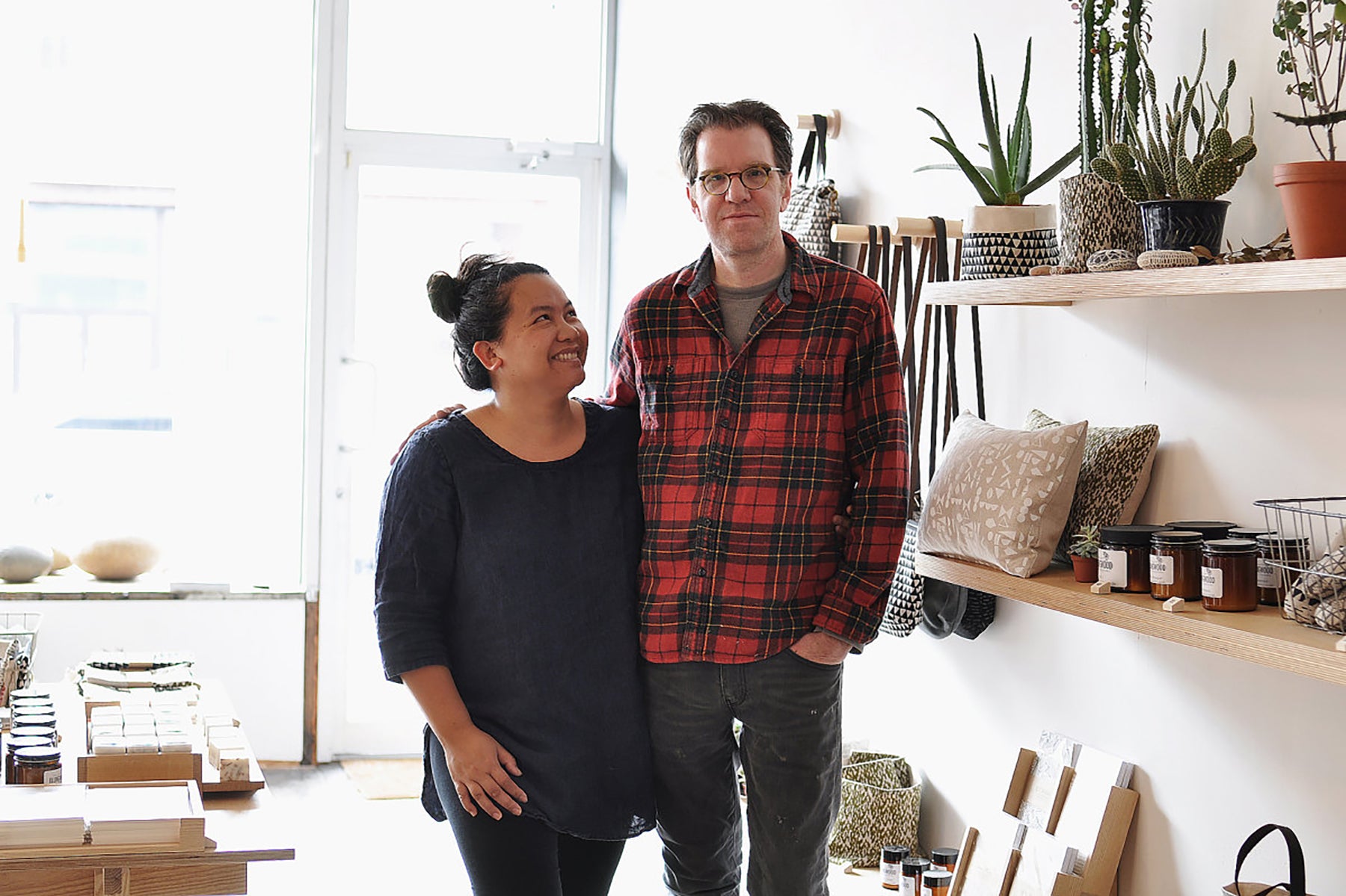 Portrait of Arounna and John, founders of Bookhou, standing in their retail store. There are handmade goods on the shelves to the right of them and Arounna is looking up a John while he looks directly to camera. Arounna is wearing a loose navy long sleeve top and John is wearing a red checkered lumber shirt. 