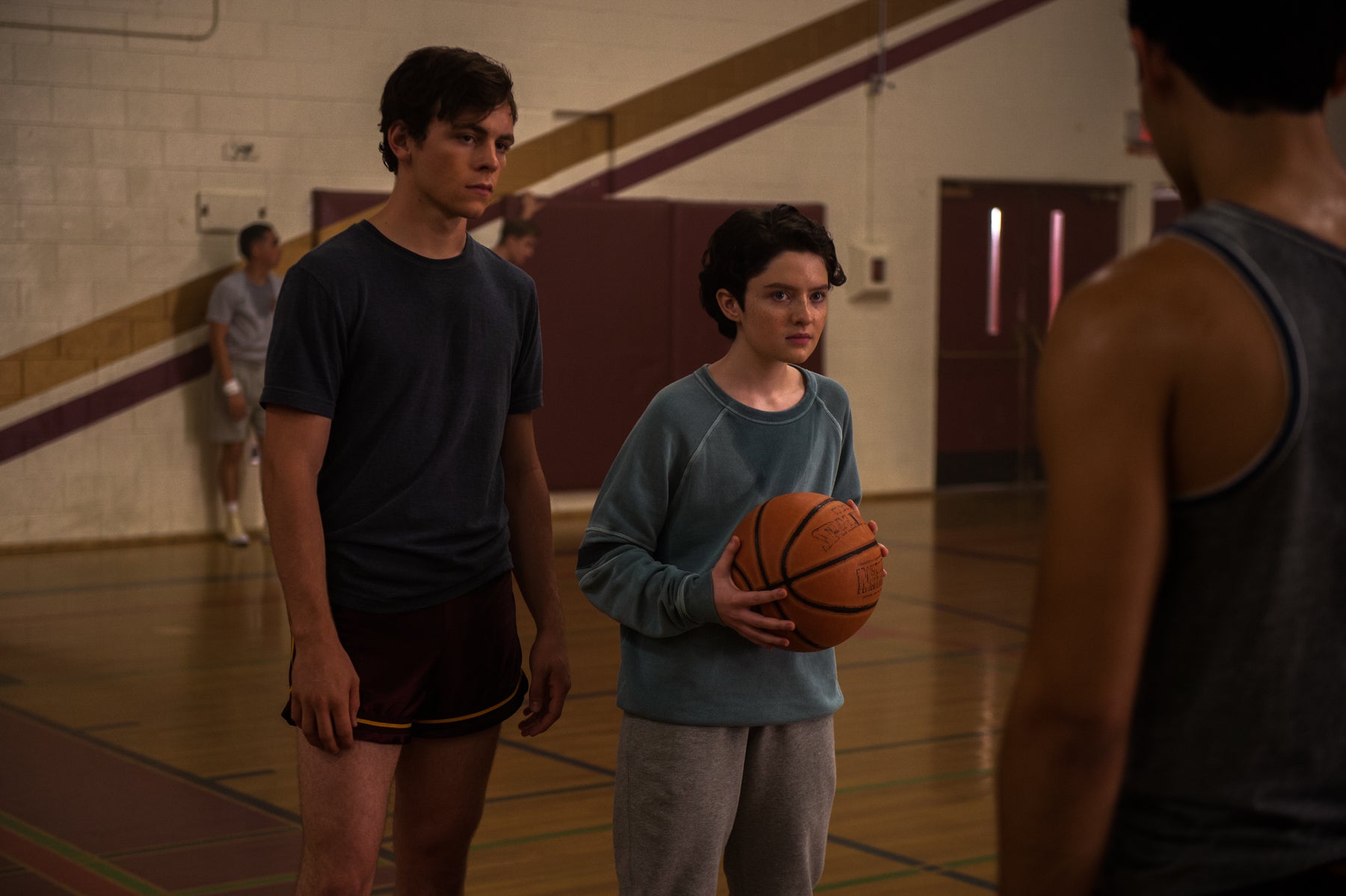 Harvey and Theo stand in the Greendale High gym, facing a character with his back turned. Theo is holding a basketball.
