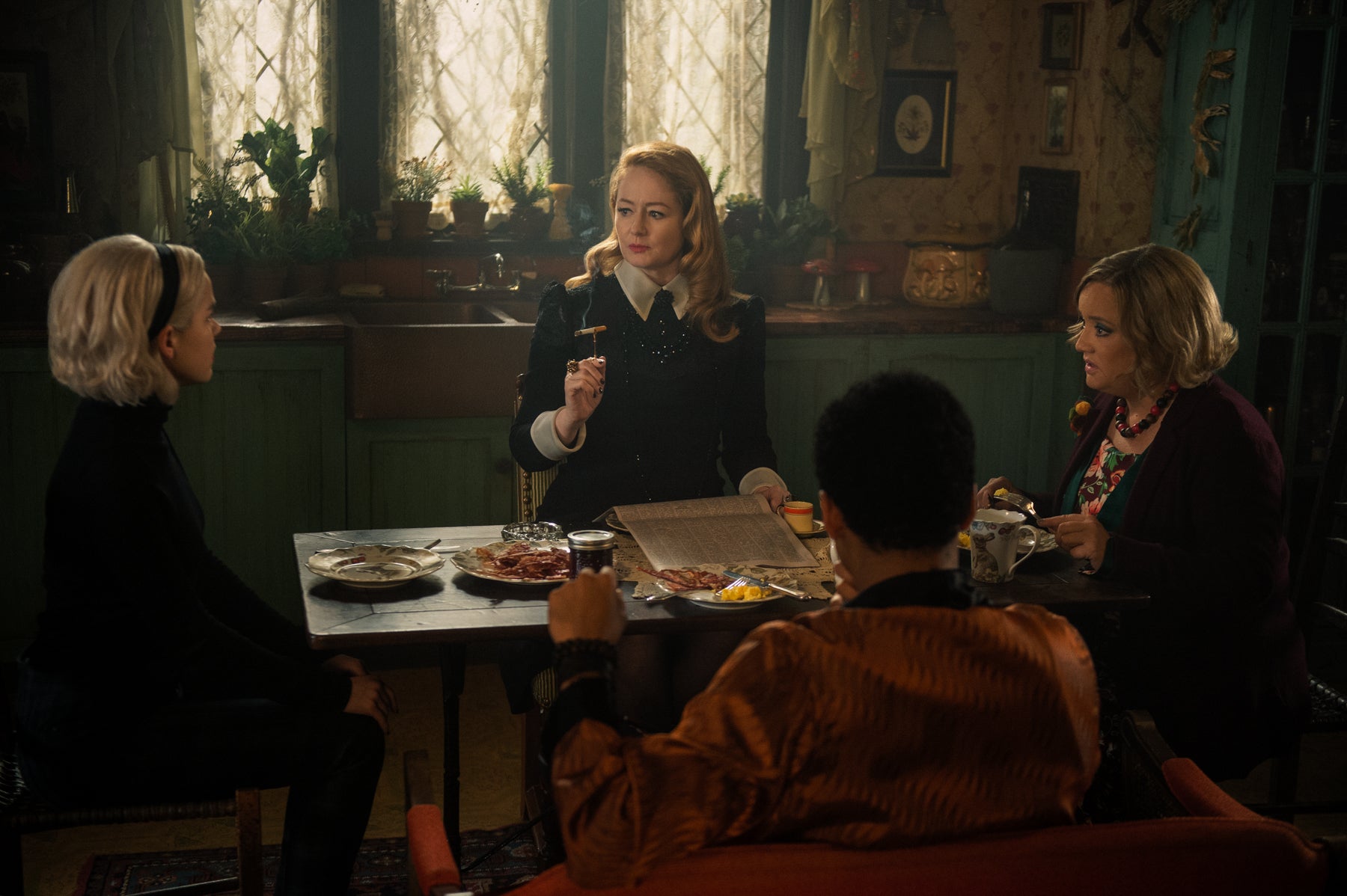 Sabrina Spellman sits at her kitchen table with her aunts Zelda and Hilda and cousin Ambrose in a scene from Chilling Adventures of Sabrina. The table is covered in breakfast plates, and Aunt Zelda is smoking a cigarette.