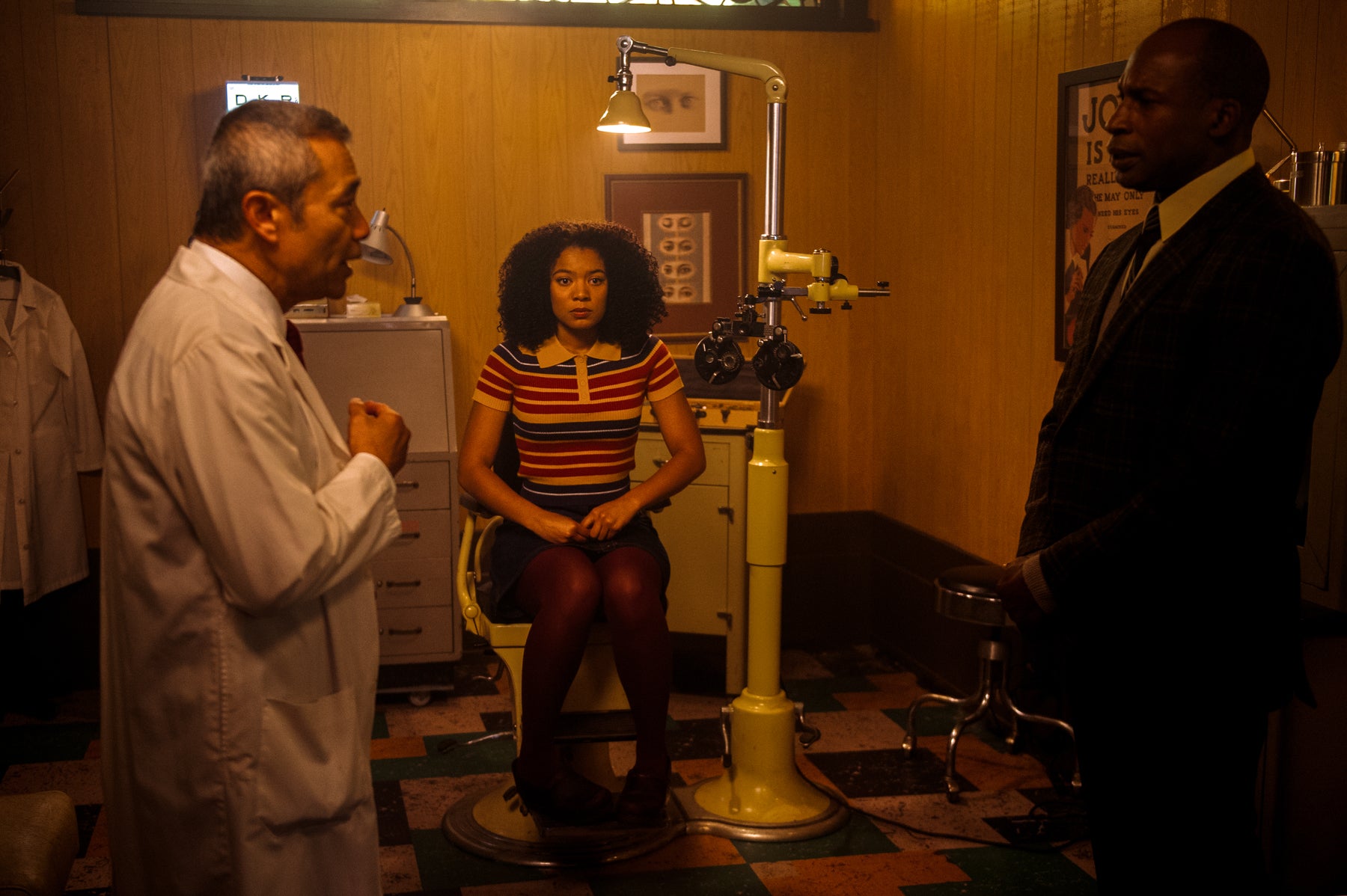 Ros sits in a chair in a medical office while an ophthalmologist speaks with her father in the foreground in a scene from Chilling Adventures of Sabrina.