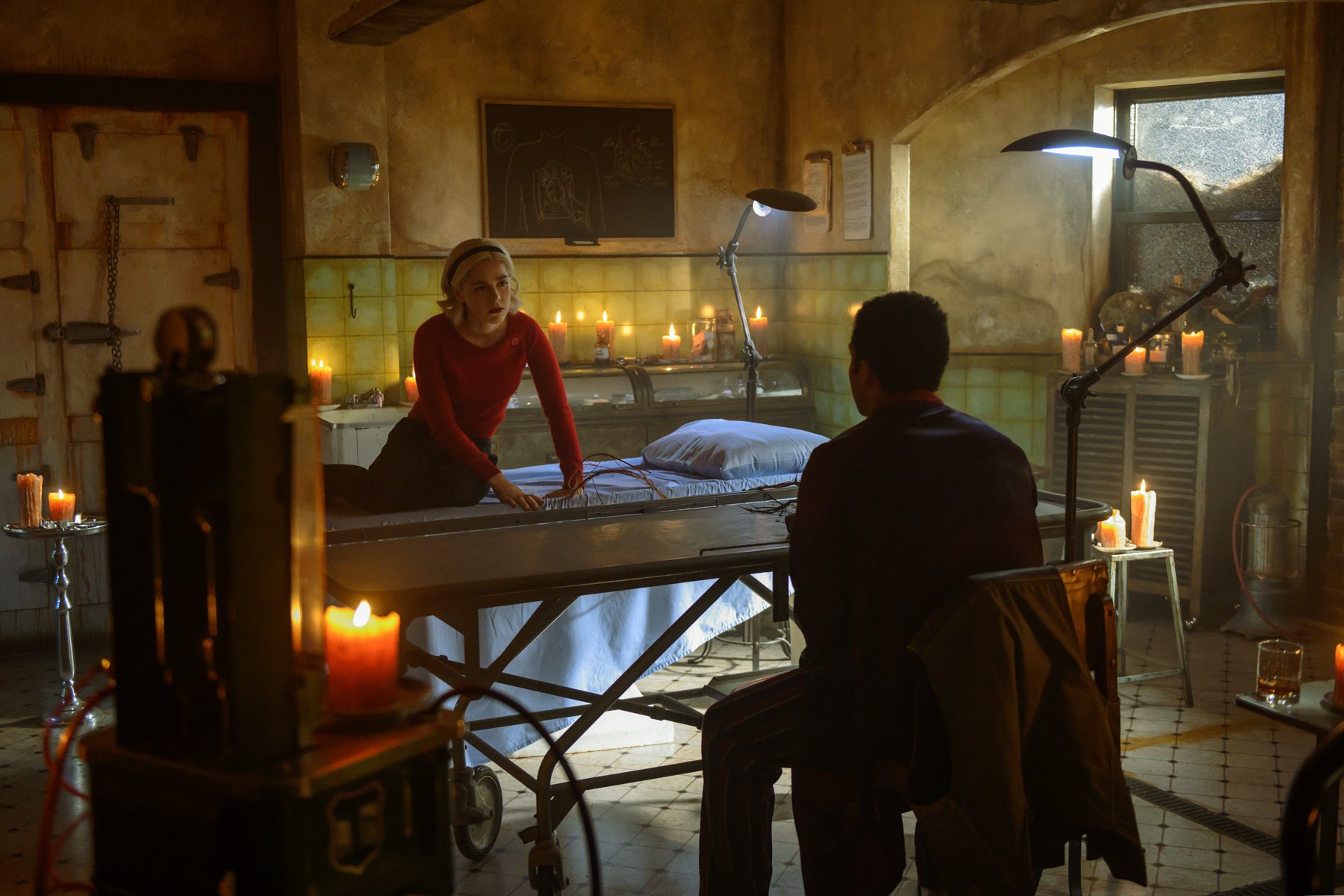 Sabrina Spellman sits on a stretcher in a mortuary speaking to a man with his back turned. The room is lit by several candles.