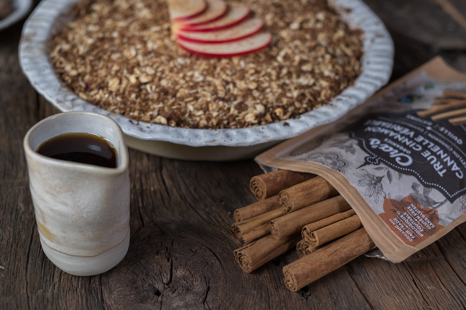 Photograph of Cha's Organics cinnamon sticks opened with a few sticks spilling out onto a wood surface. Beside it is a small vessel of maple syrup and a bowl of hot cereal in the background, slightly out of focus.  