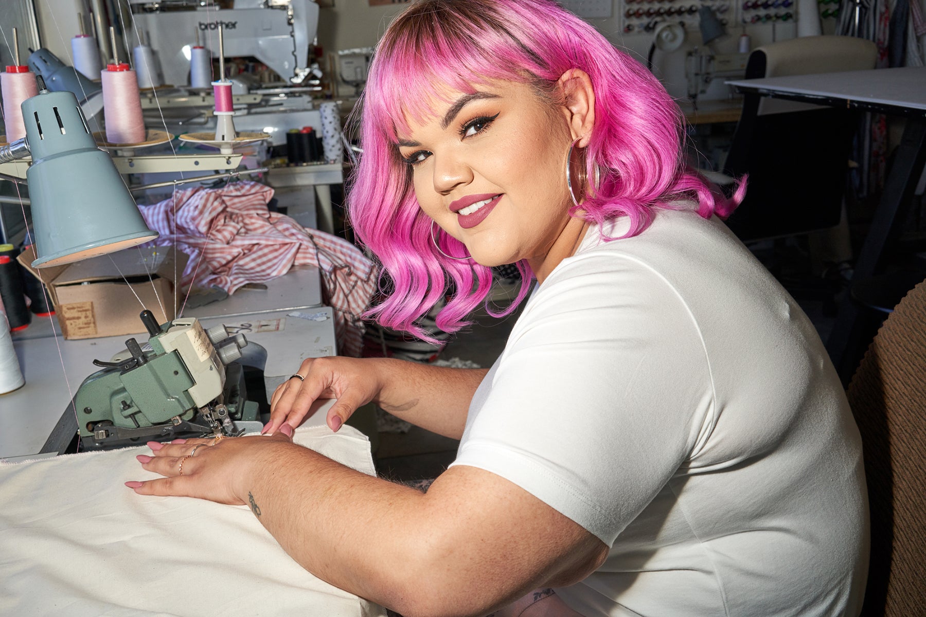 Plus fashion designer Ashley Nell Tipton with vibrant pink hair, smiling and sewing fabric.