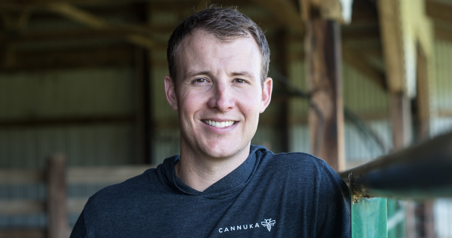 Portrait of Cannuka CEO Michael Bumgarner, standing inside his barn smiling at camera wearing a black Cannuka sweater. This is a tight portrait of his face and the background is out of focus. 