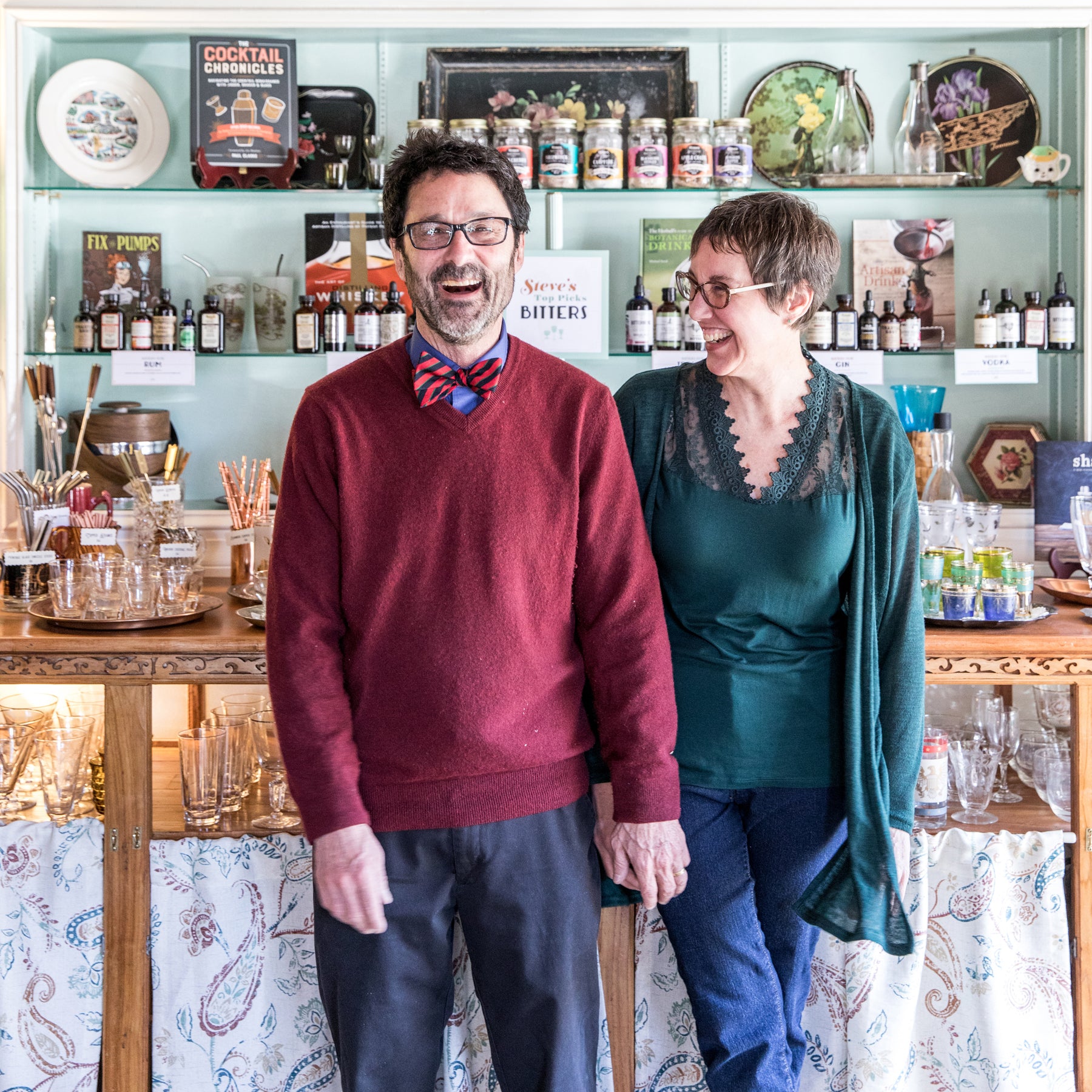 A man and a woman stand holding hands and laughing in front of a retail display of cocktail bitters and glassware.