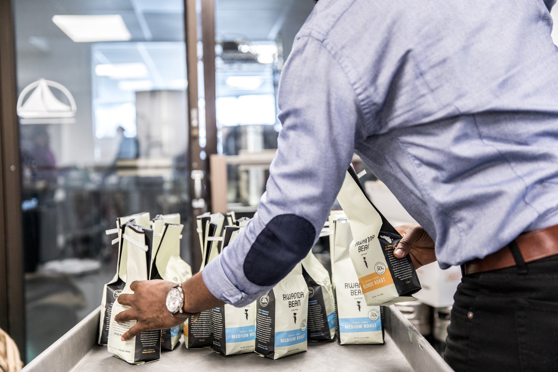 Detail of a man stacking bags of coffee beans on a table in a production facility. The bags are labeled “Rwanda Bean.”