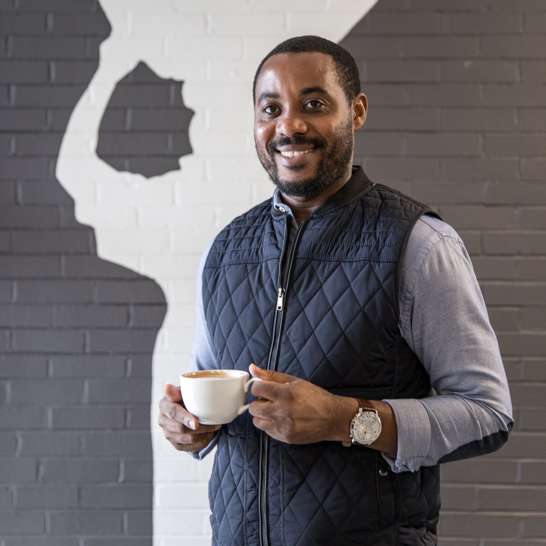 Portrait of Mike Mwenedata, founder of Rwanda Bean, holding a cup of coffee and standing in front of a brick wall. 