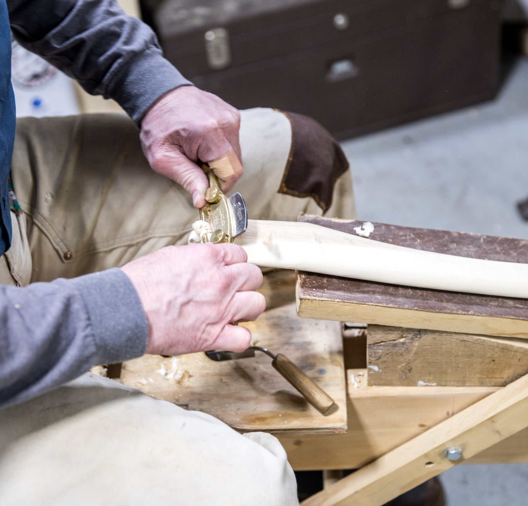 Detail of a man’s hands working with a metal tool on a wooden axe handle.