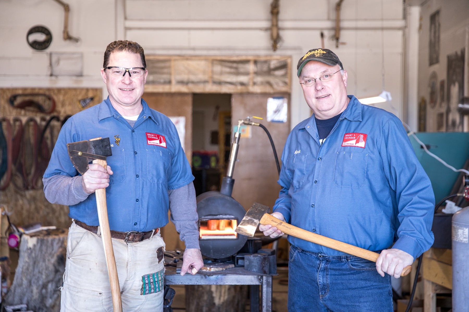 Brant & Cochran founders Barry Worthing and Mark Ferguson stand holding axes in their Portland, Maine workshop. 