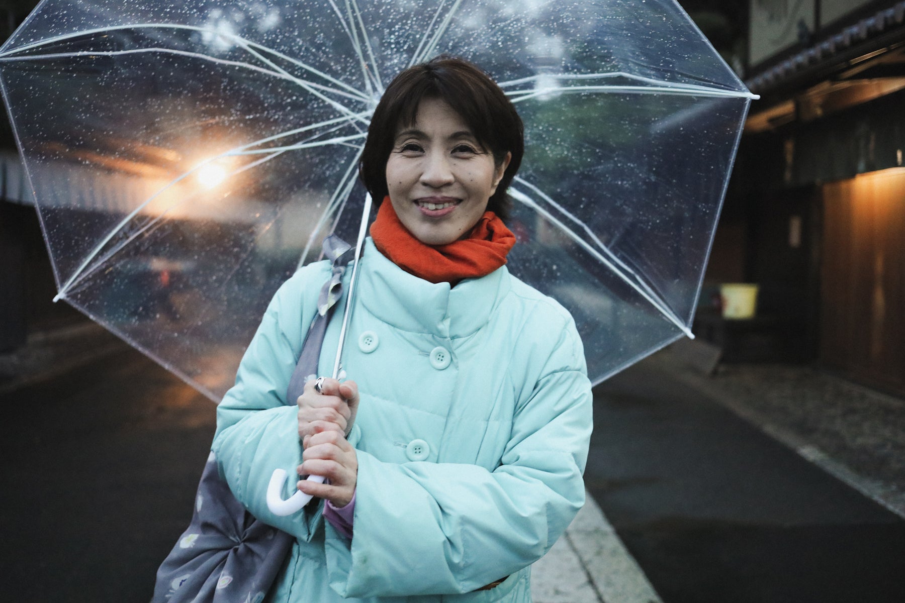 Etsuko Yamada, owner of Musubi Furoshiki, poses in a pale blue jacket and red scarf with a transparent umbrella on the rainy streets of Kyoto. 