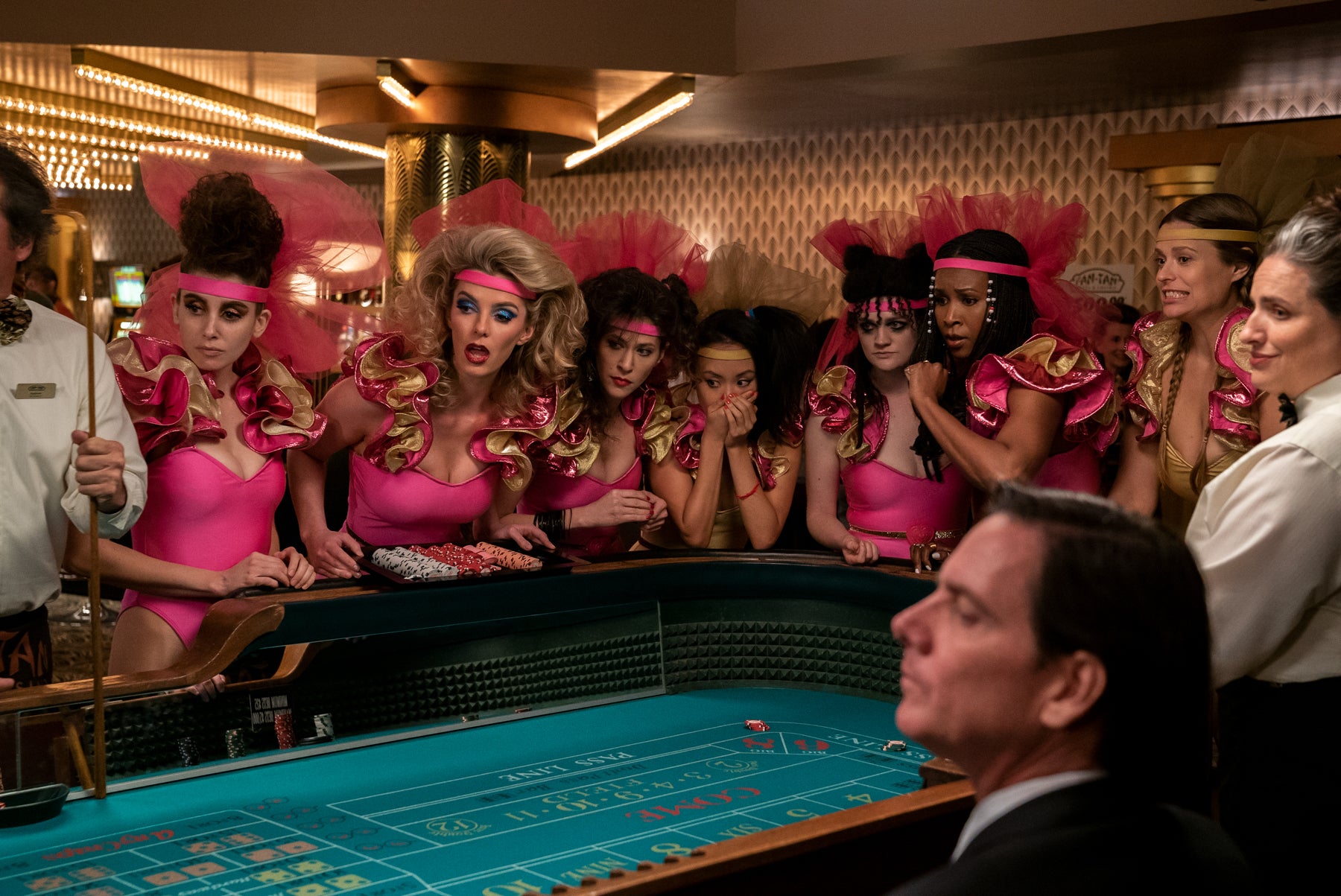 A photo from Netflix’s GLOW with wrestlers gathered around a craps table in Vegas while dressed in frilly pink-and-gold stage costumes. Cherry Bang had just rolled the dice, and they’re all watching in anticipation to see if she won. 