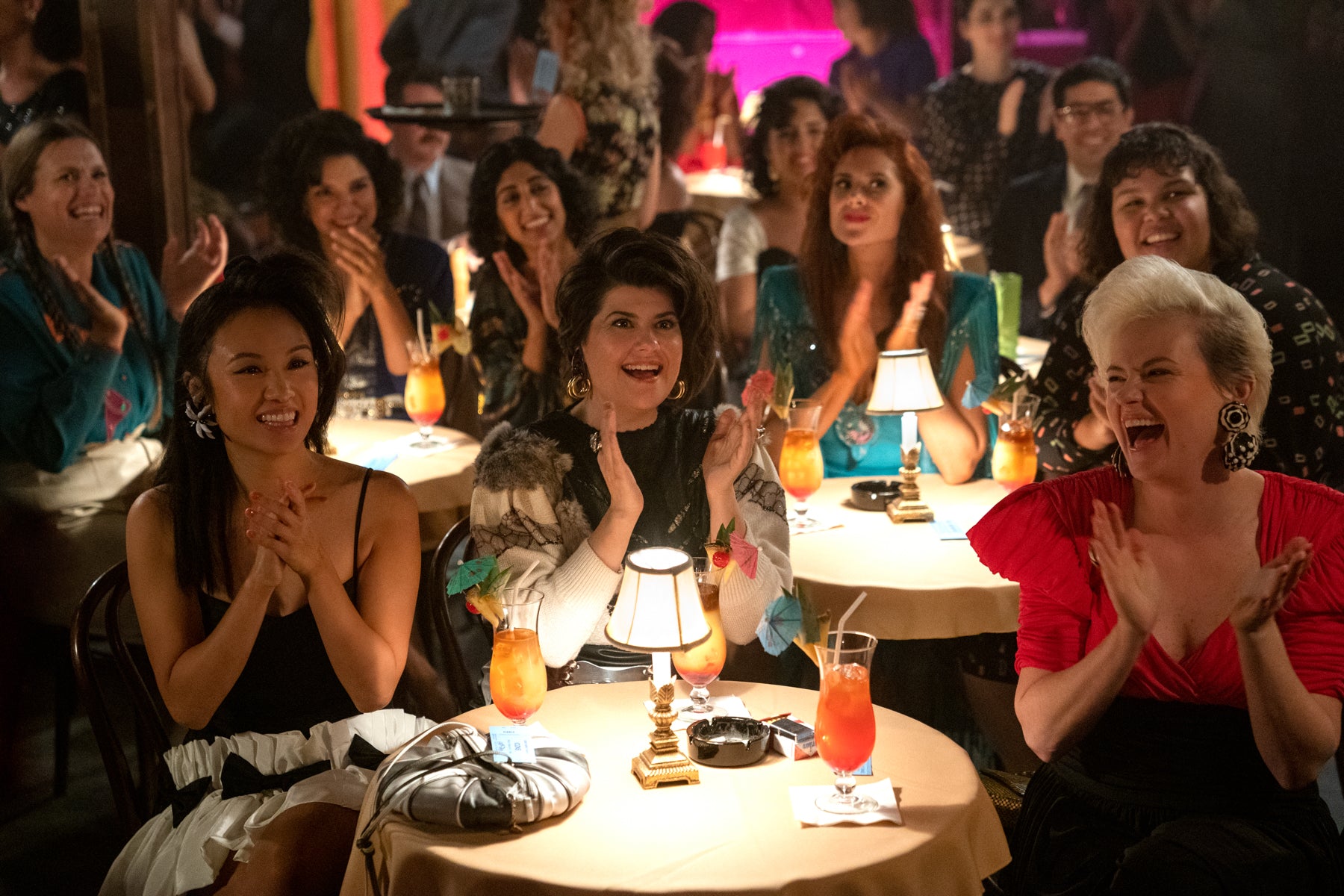 The entire cast of GLOW applauds while watching a dinner show in Vegas. They’re all smiling and laughing. 
