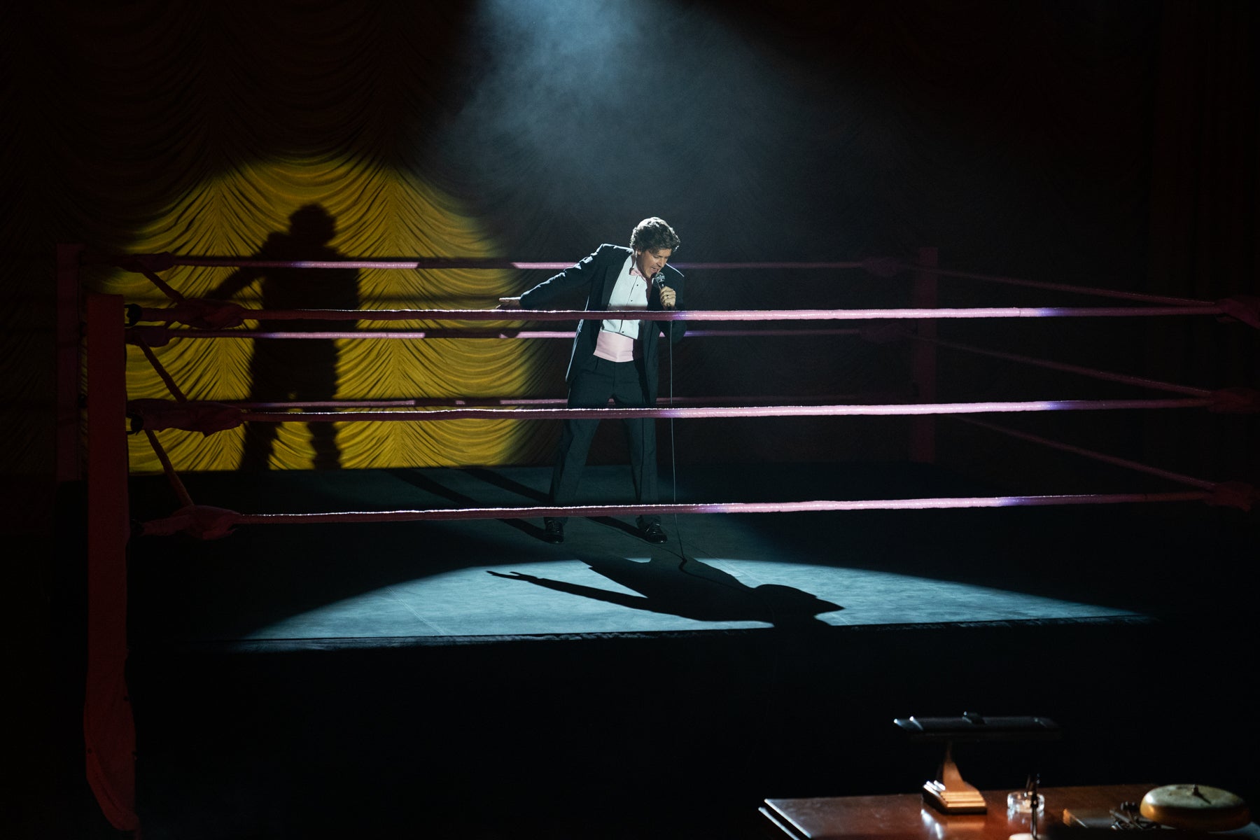 Host/announcer Bash stands in the spotlight in the middle of the wrestling ring doing the intro for GLOW, during a scene from Season 3.  