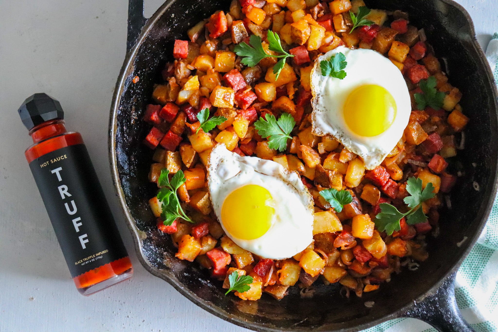 Hash browns in an iron skillet topped with two sunny side-up eggs and a bottle of Truff sauce on the side. 