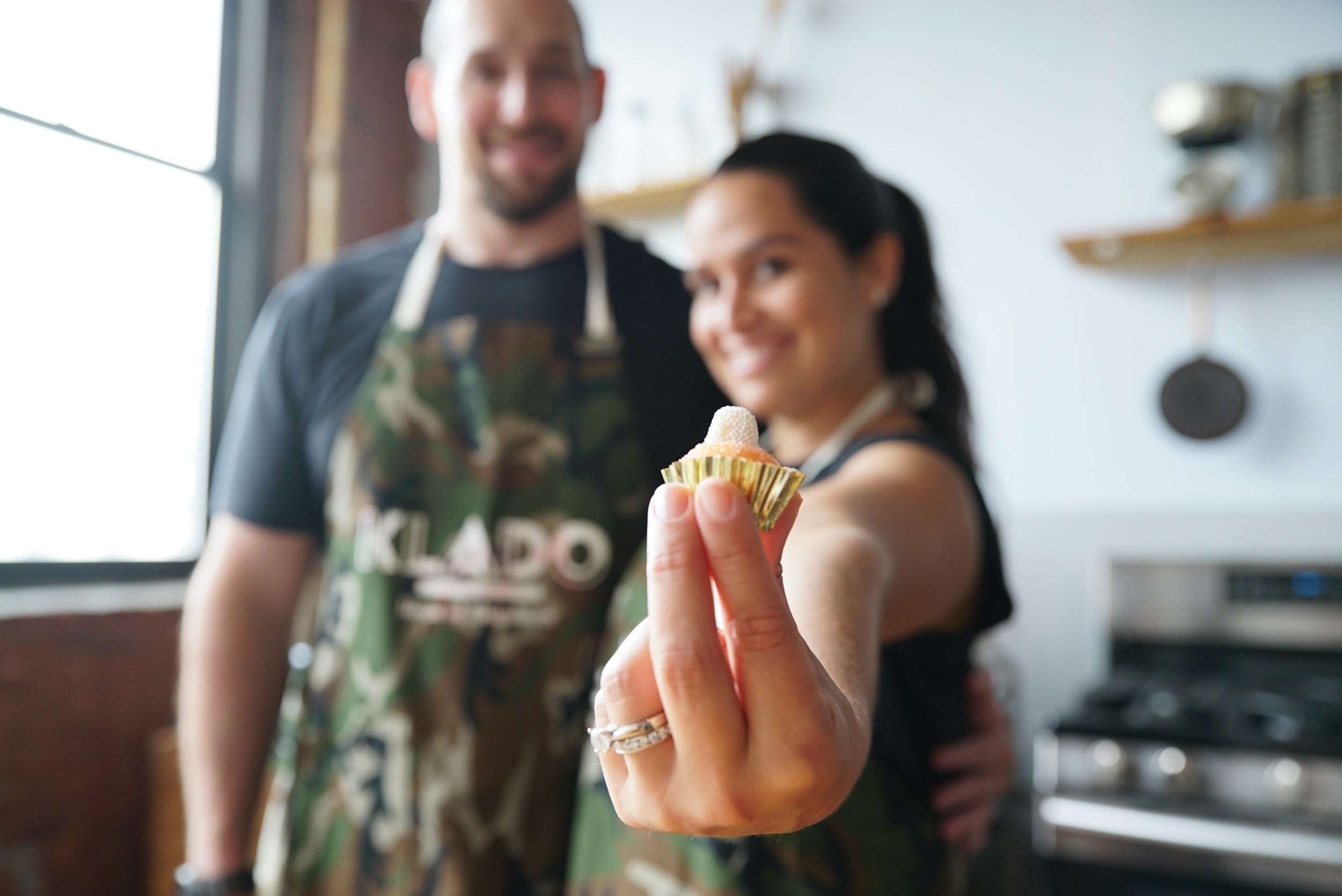 Two people stand, out of focus, wearing aprons. The woman on the right holds out a dessert, in focus in the foreground 