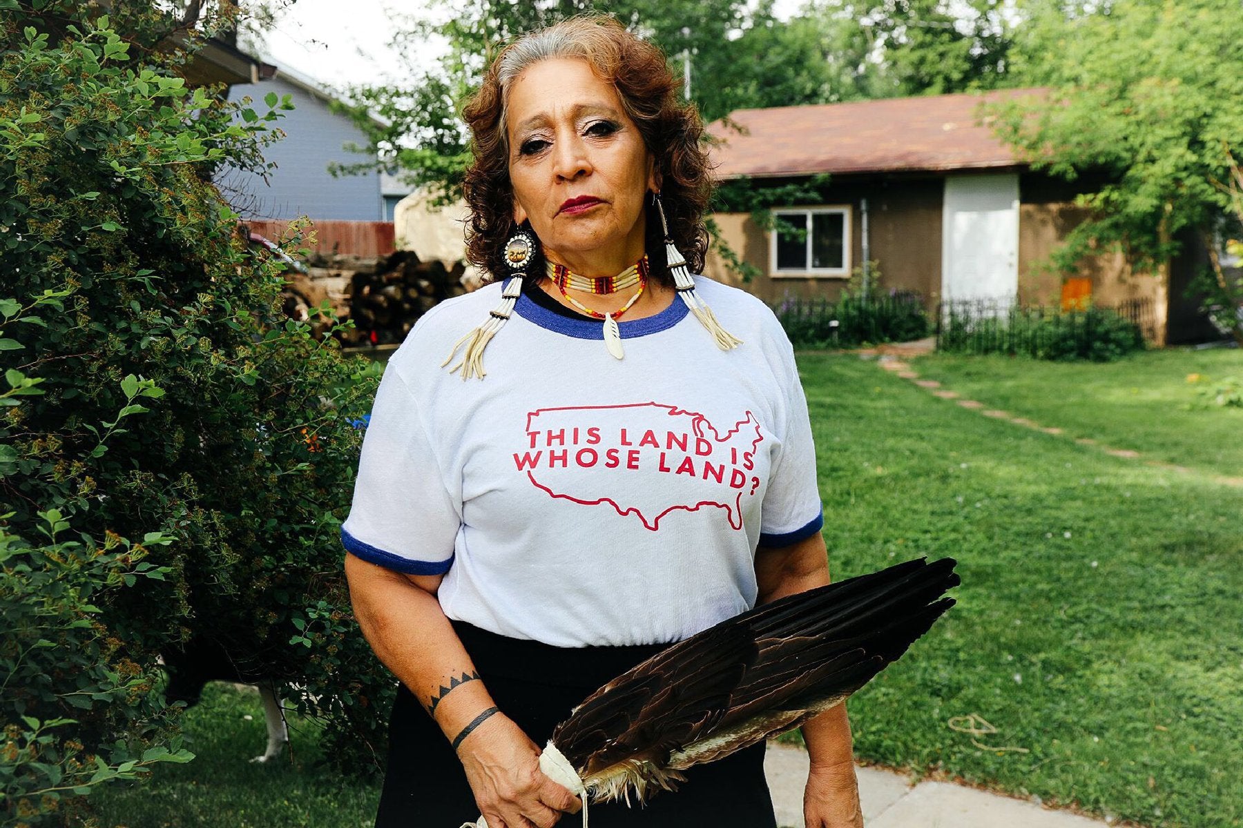 An Indigenous woman stands in a yard wearing a shirt that reads, "This land is whose land?"