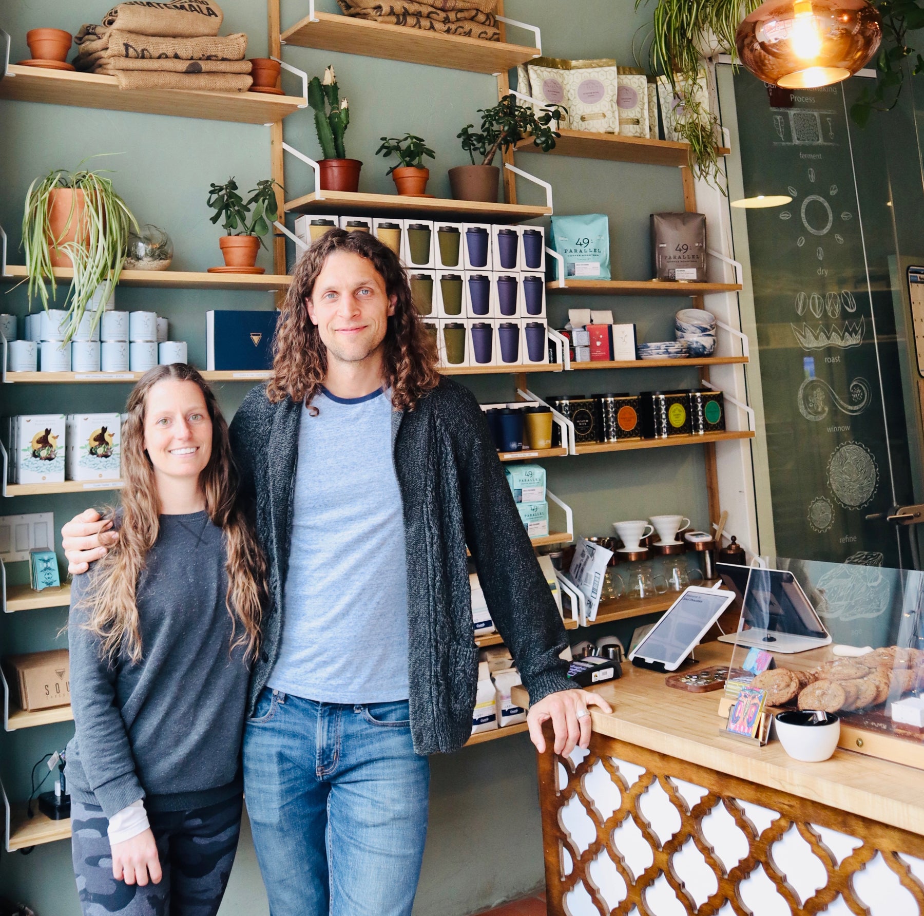 Katie Wilson is on the left and in a gray shirt with camouflage pants. Kyle Wilson is on the left in gray T-shirt, dark gray hoodie, and jeans. The couple and business partners are standing in front of the shelves displayed in their shop, Soul Roasters. 