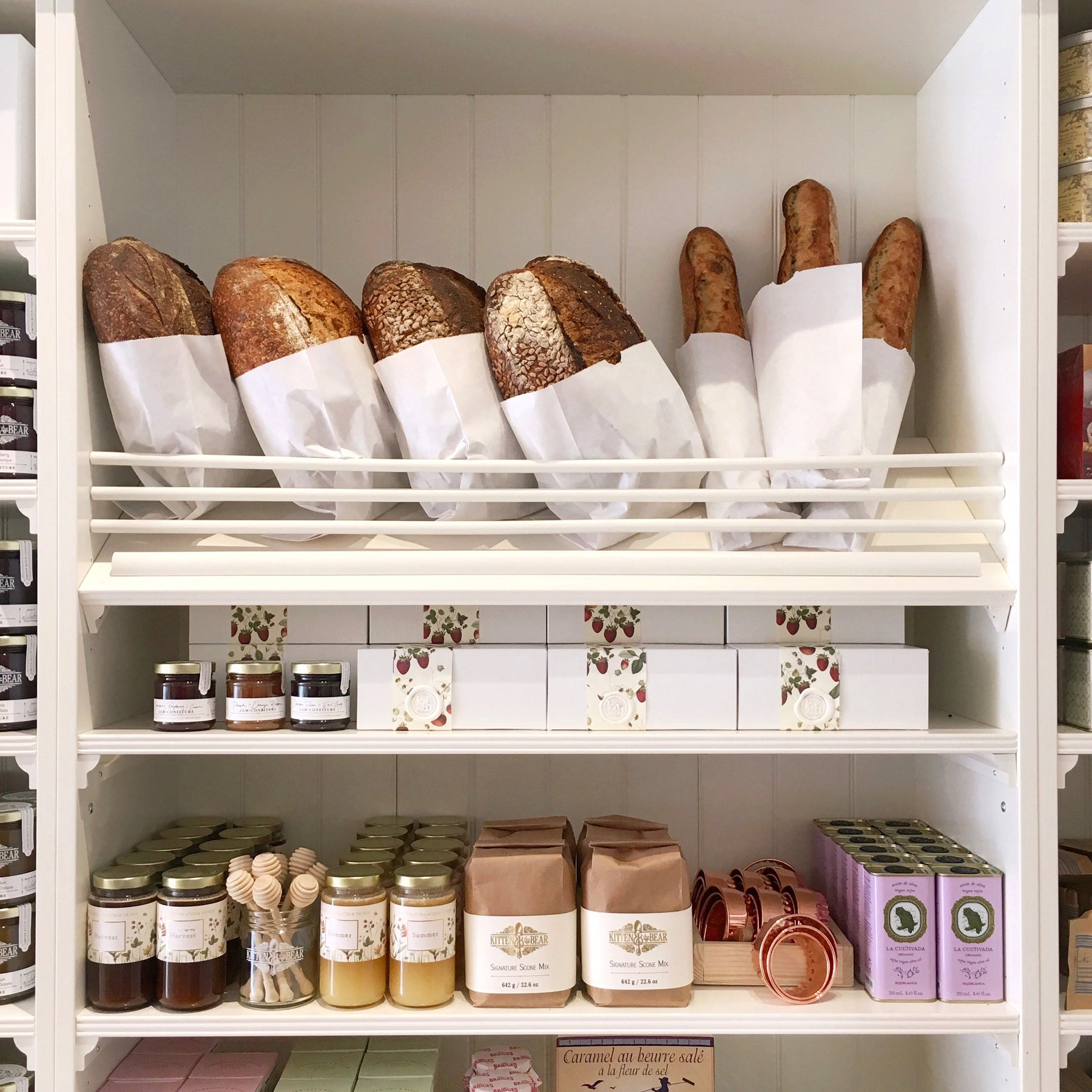 White wooden shelves within Kitten and the Bear hold fresh baked bread (top shelf), seasonal preserves (middle shelf), and pre-packaged baking mixes and baking ware (bottom shelf).