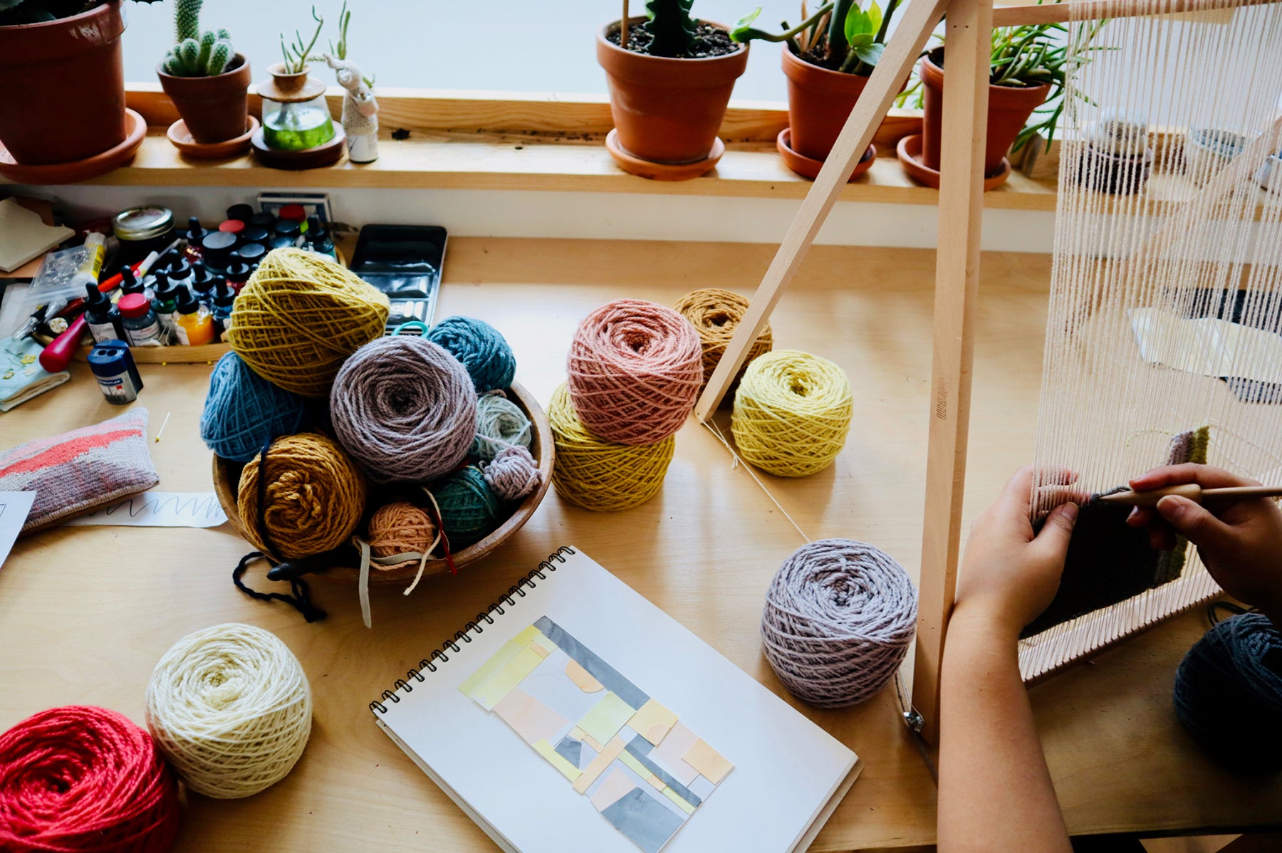 Still life photograph of spools of different coloured spools of yarn, spilling out on a table. Arounna's hands are pictured working on her loon and the reference artwork is resting on the table in front of her.