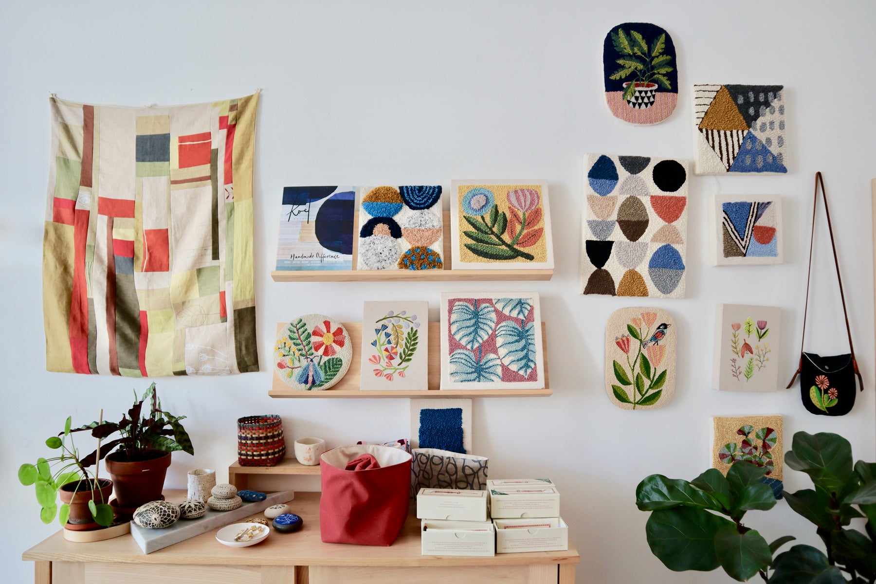 On the wall of Arounna’s shop hangs a patchwork quilt (left), a collection of 13 punch needle on canvas pieces (center), and a shoulder bag with a punch needle design (right). In front of the wall is a table with plants, rockets covered by crochet, project bags, and punch needles in boxes. 