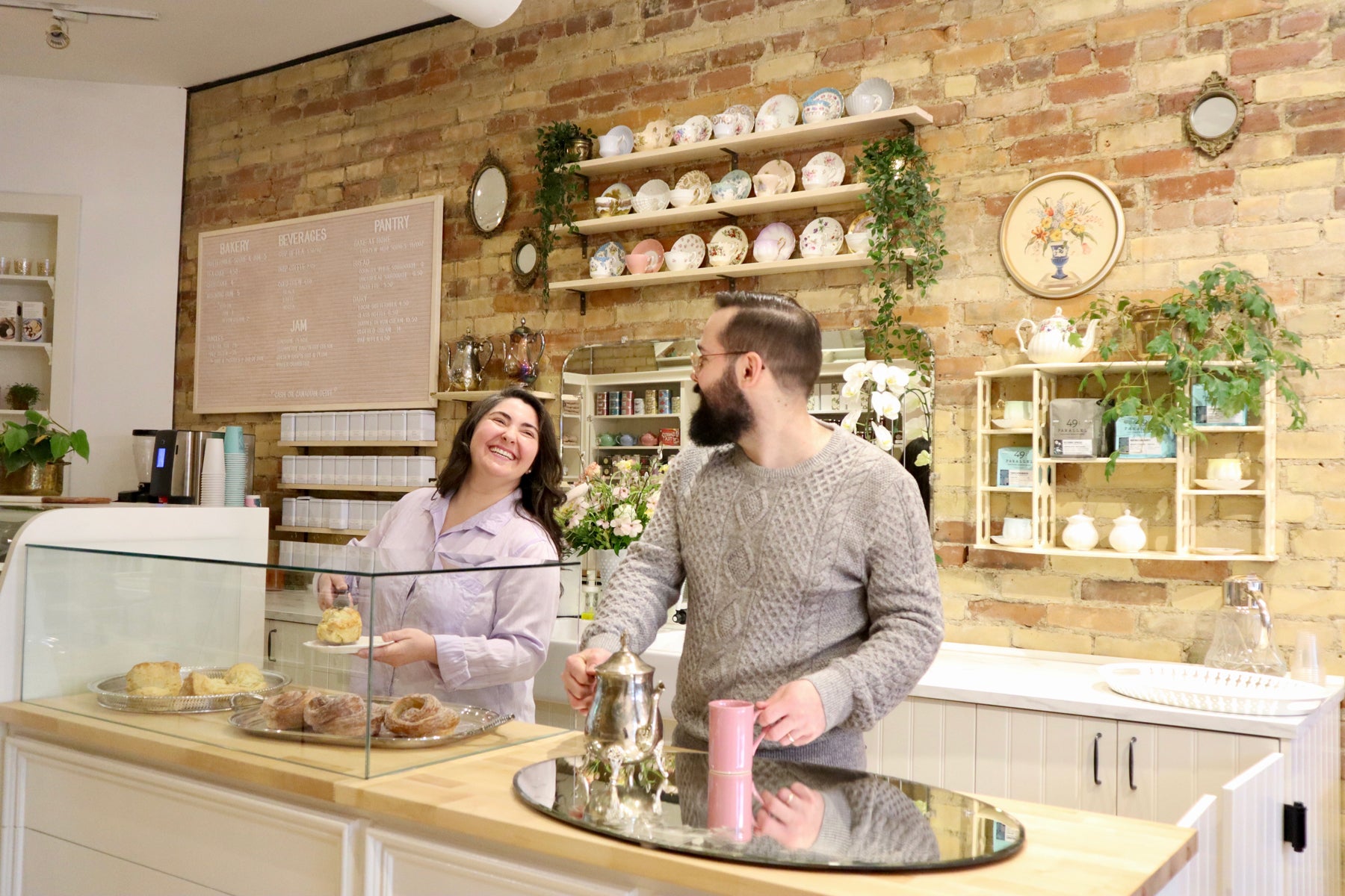 Sophie Kaftal, in a purple shirt, plates a freshly baked scone in the new Kitten and the Bear location. Meanwhile, her husband and business partner, Bobby Zielinski, wearing a gray sweater, preps a hot beverage. 