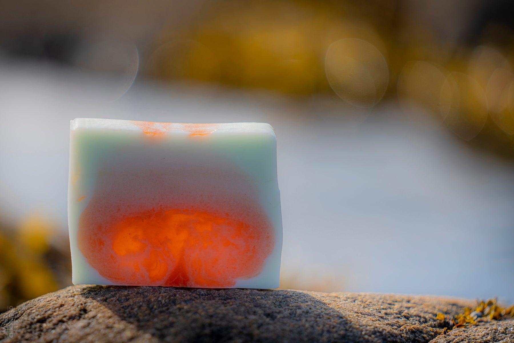 Close up of a bar of UasaU soap sitting on a rock. The soap is glowing as it is back lit by the sun. The soap is a mottled orange and white.