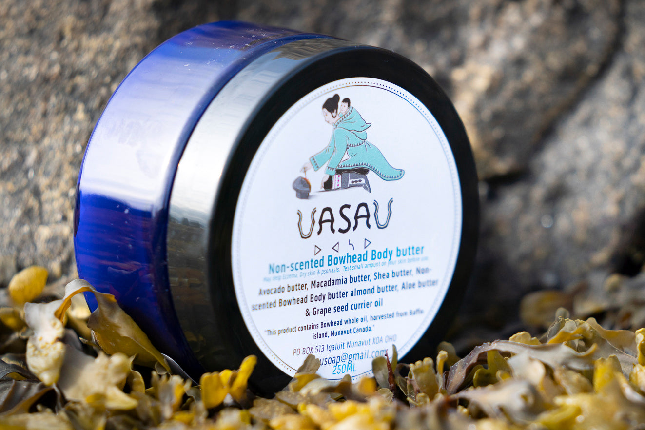Close up of a circular jar of UasaU soap body butter placed on a rock with lichen surrounding it. The label features the UasaU soap logo which is a drawing of an Inuit mother holding her child. 