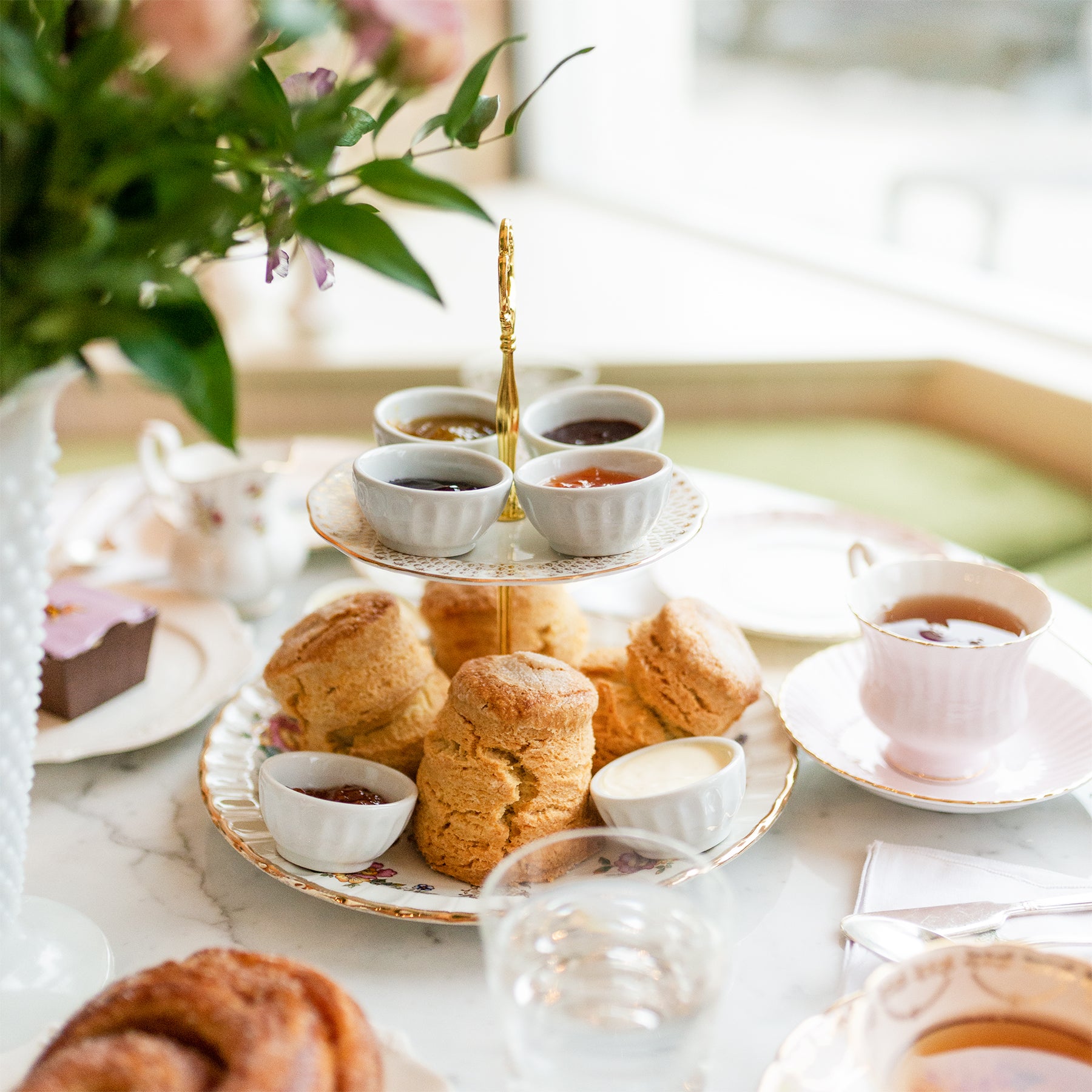 The tea service scene at Kitten and the Bear shows a table, with a two-tiered display. There are four preserves on the top tier and four scones on the bottom tier along with another preserve and clotted cream in ramekins. 