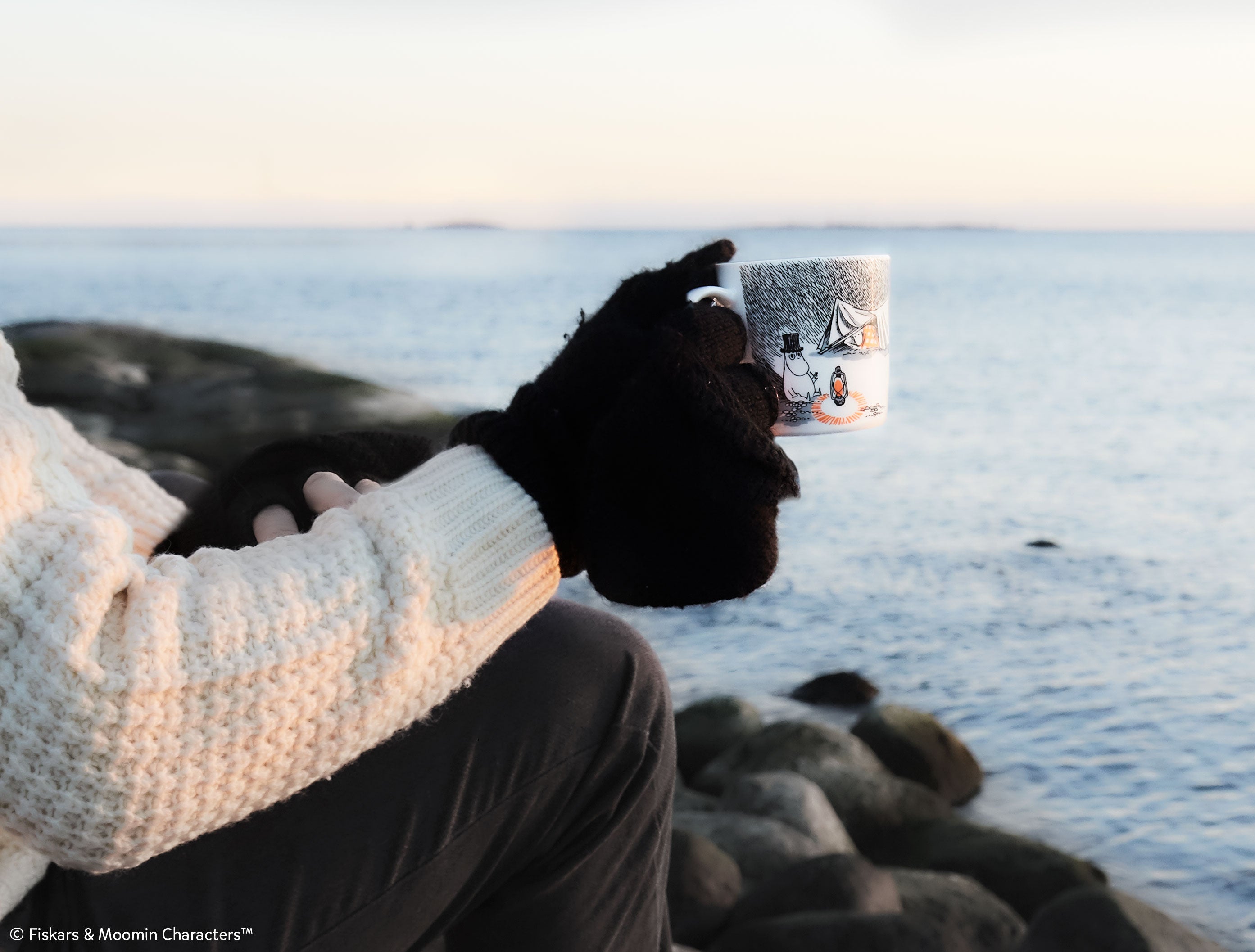 A Moomin cup held by a hand model, backdropped by the ocean. 