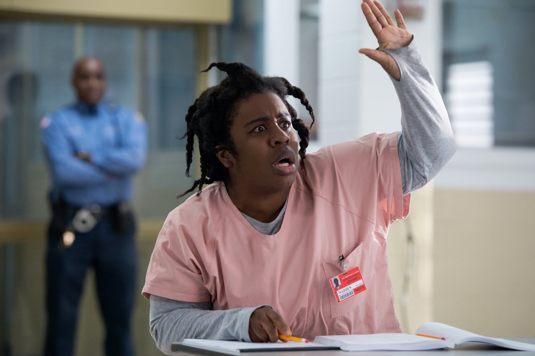Suzanne Warren sits at a desk raising her hand while a prison guard stands in the background in an episode of Orange Is the New Black.