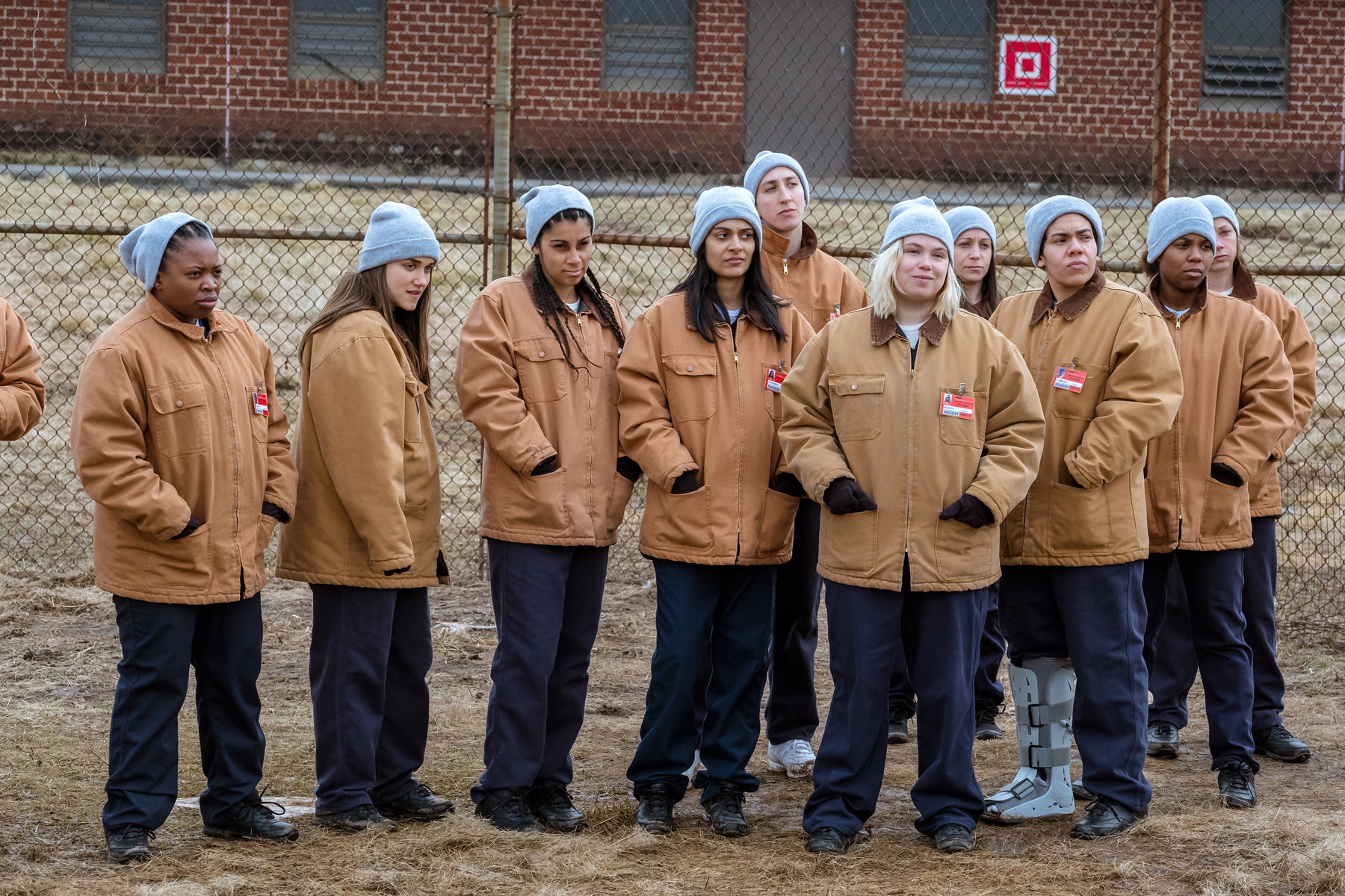 Inmates in coats and hats stand in a line against a prison fence in an episode of Orange Is the New Black.